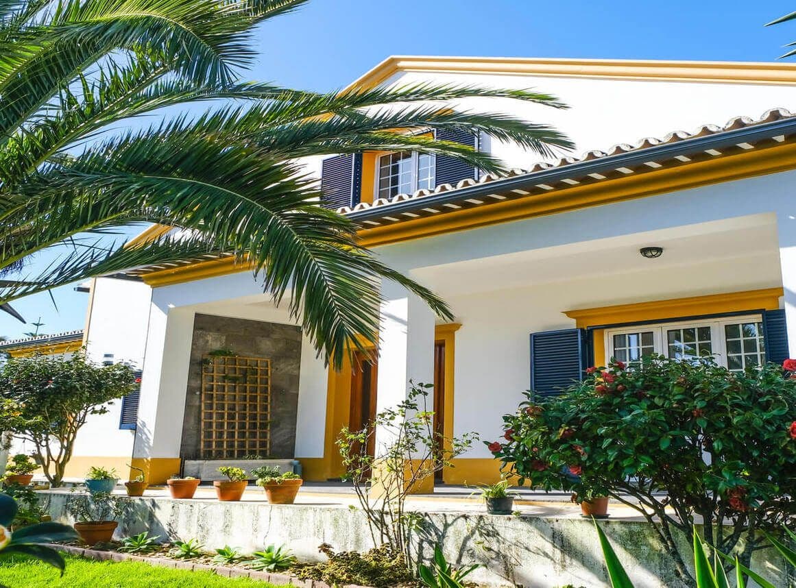 Front exterior view of the Lava Coliving house with white and yellow facade, palm trees and tropical garden