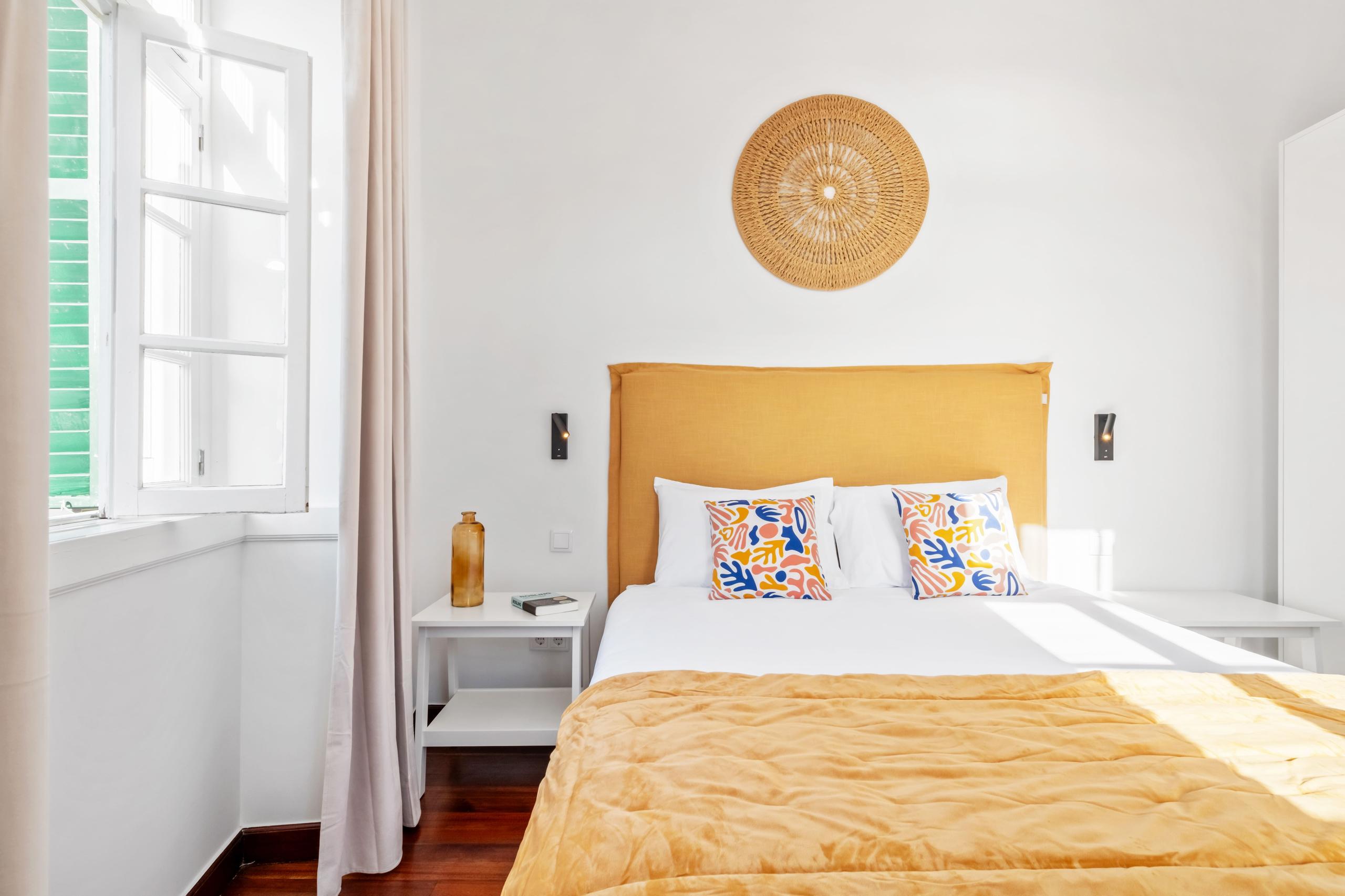 Spacious room with queen bed, yellow headboard, colorful cushions, and desk with monitor