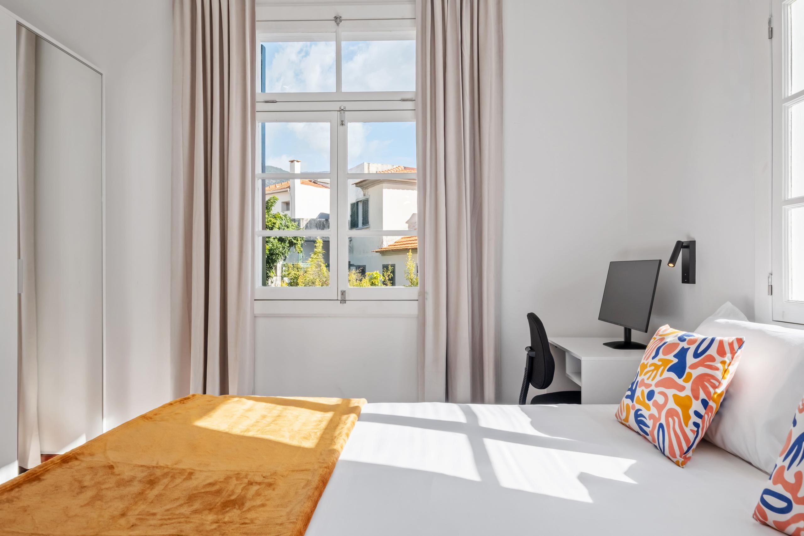 Well-proportioned room with queen bed, colorful cushions, desk with monitor, and two windows