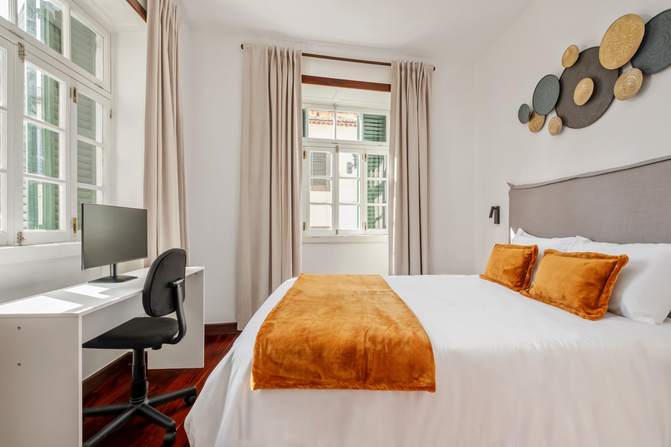 Strelitzia room showing queen bed with orange cushions, desk with monitor, and windows with green shutters
