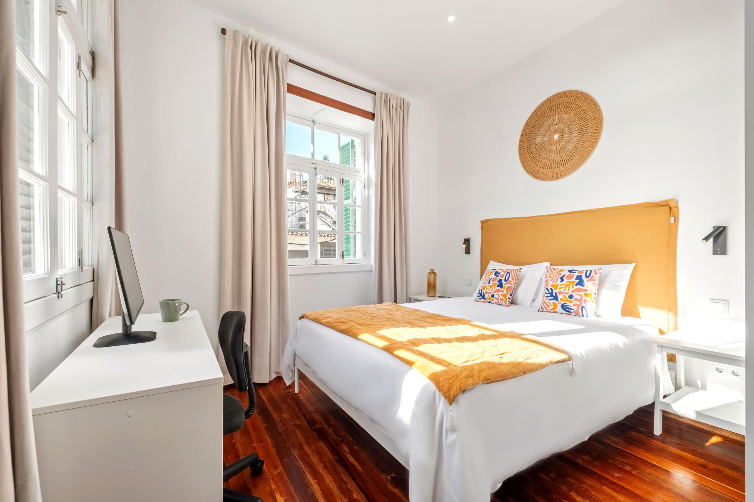 Hortênsia room showing queen bed with yellow headboard, desk with monitor, and windows