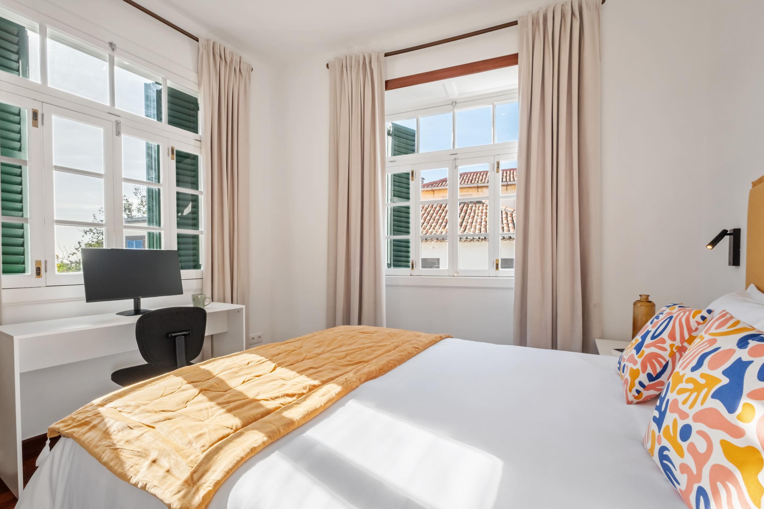 Hortênsia room wide angle showing queen bed, two windows with green shutters, and desk area