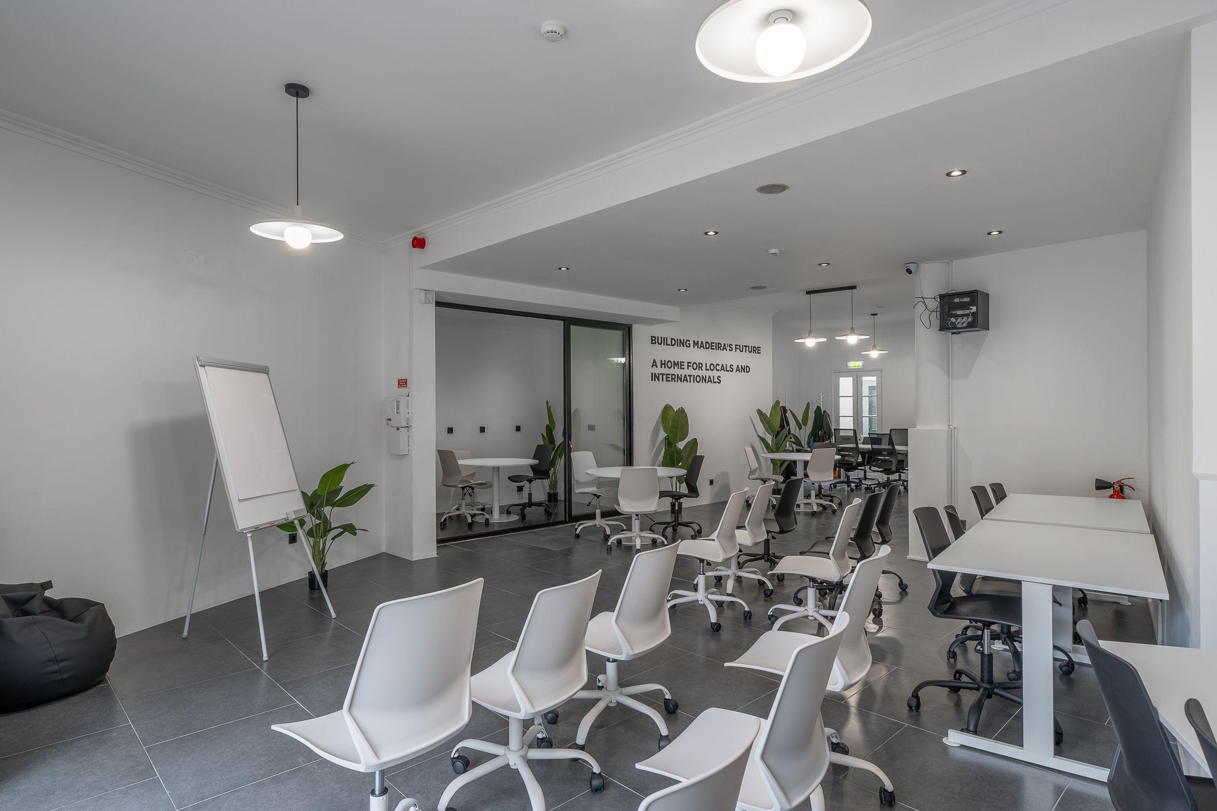 Main coworking area with rows of white chairs and desks, whiteboard, plants, and wall text reading 'Building Madeira's Future'