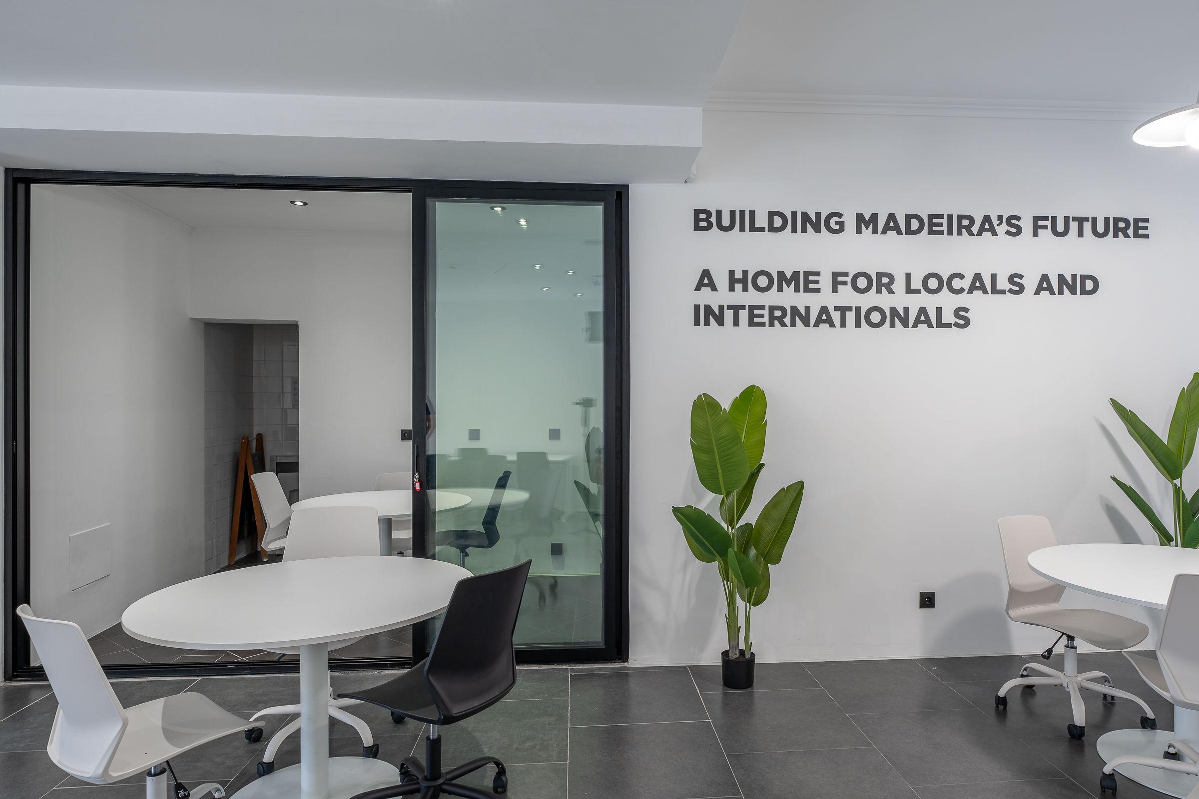 Coworking space showing glass-walled meeting room, round table, and wall text 'Building Madeira's Future'