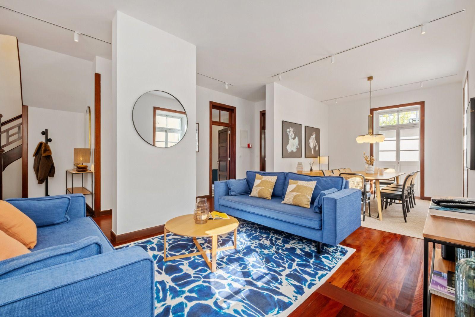 Spacious living room with blue sofas, round mirror, dining area, and wooden floors