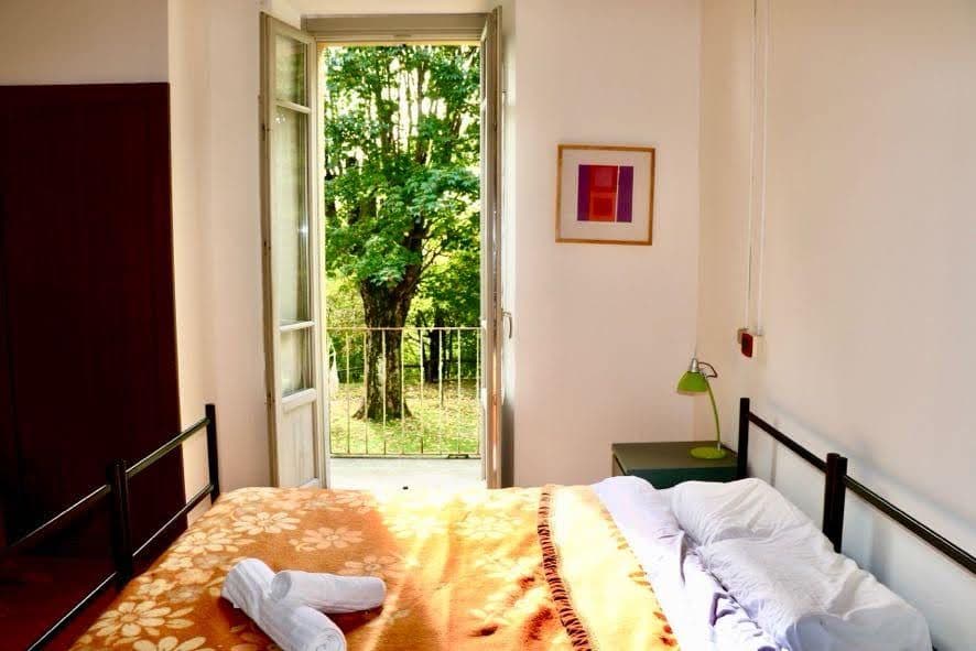 Bright private double room with open balcony door overlooking green trees, a bed with orange floral bedspread, and a side table with lamp