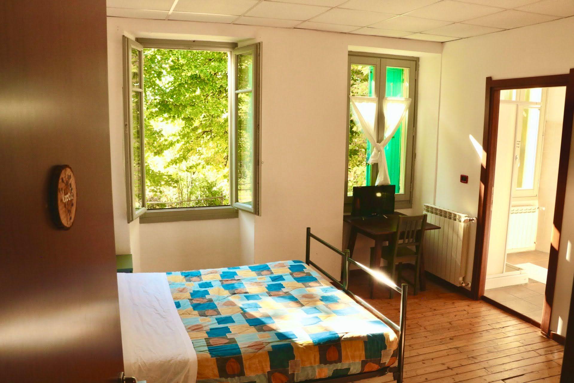 Bright private double room with two open windows overlooking green trees, a desk with laptop, and a double bed with colorful quilt
