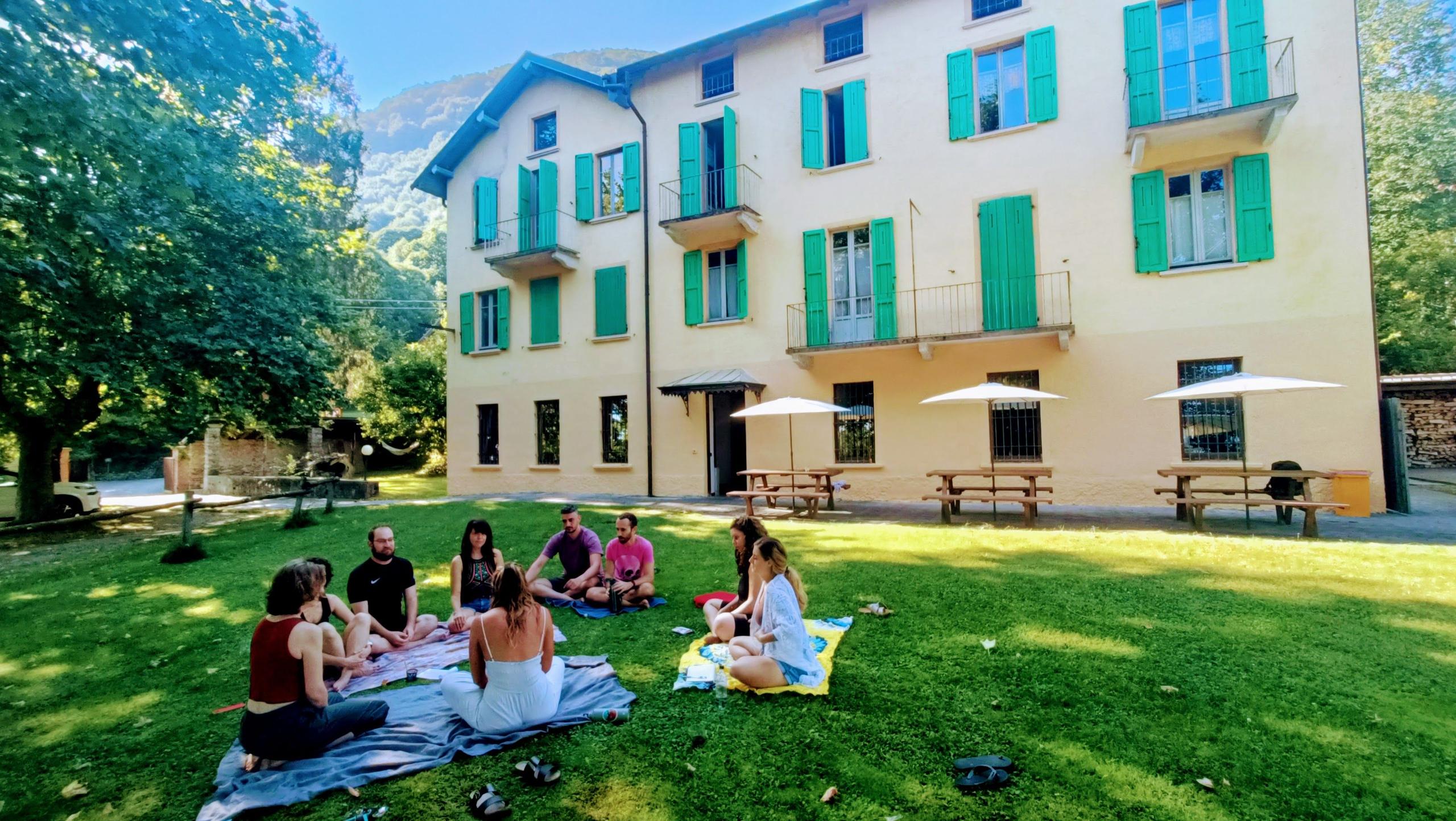 Group of residents sitting in a circle on the lawn in front of the Montino building, with the building facade and green shutters visible in the background