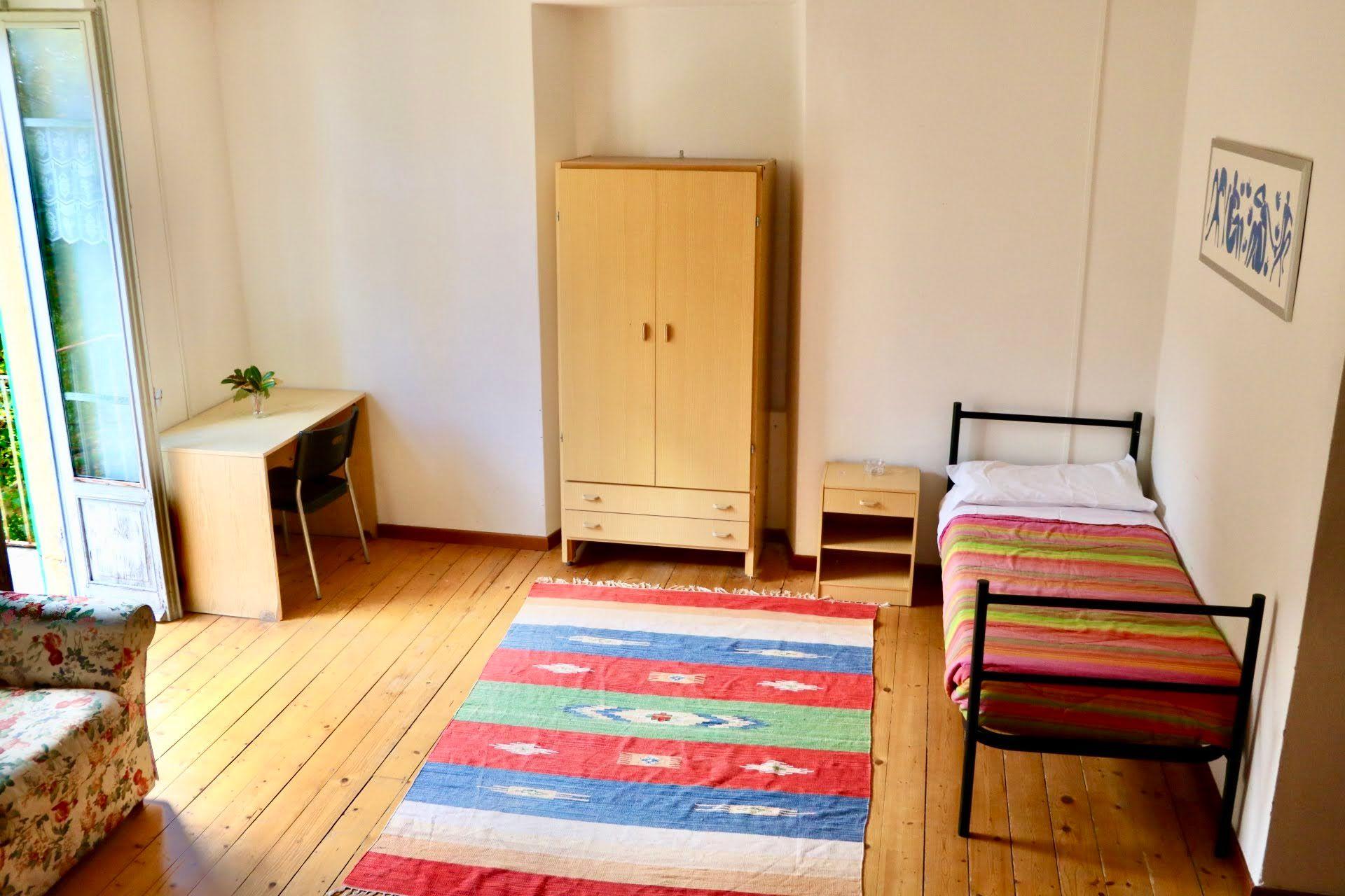 Bright single private room with a single bed, wardrobe, desk, and colorful rug on wooden floors