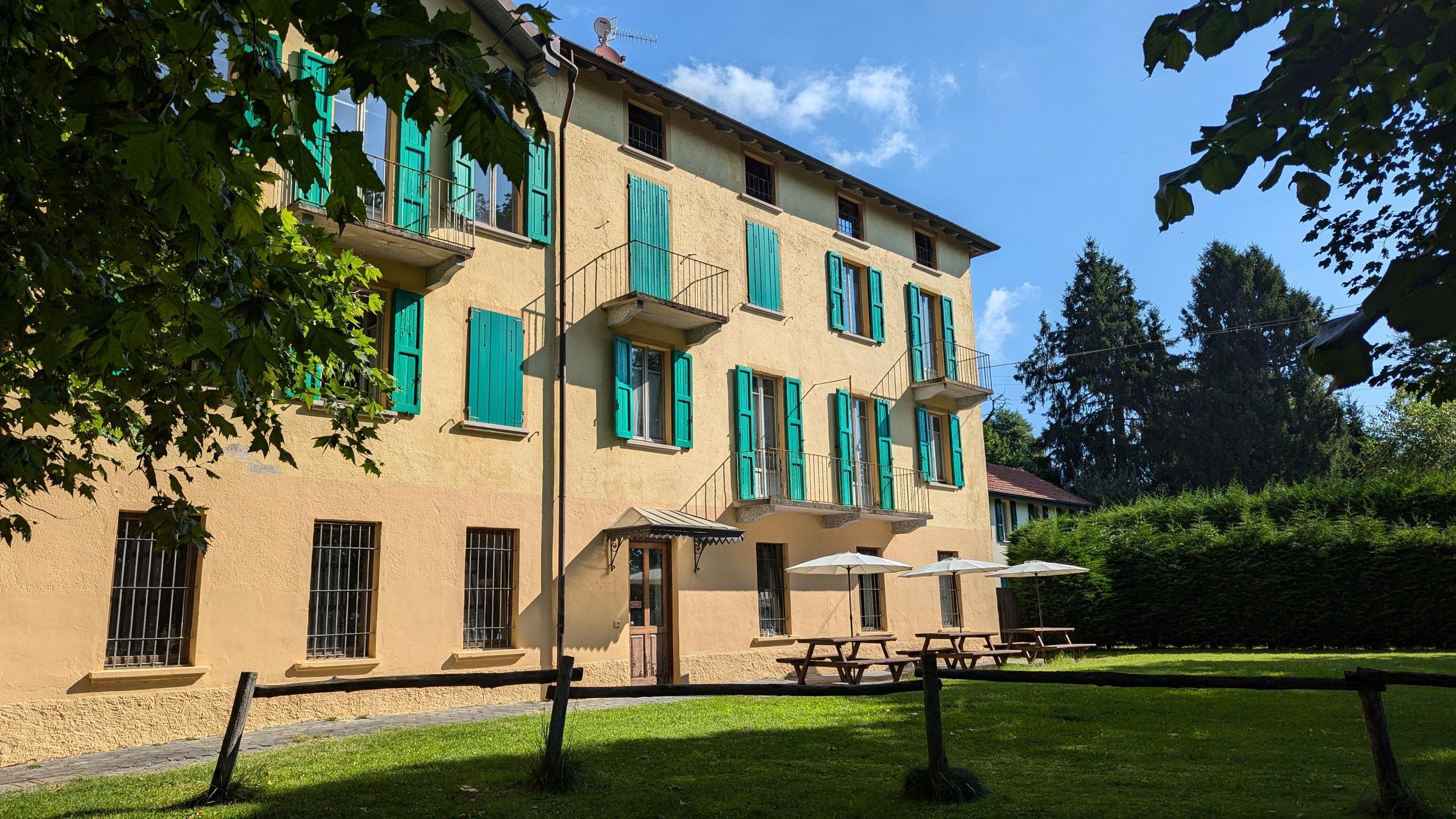 Exterior view of the Montino building with green shutters and balconies, seen from the garden with trees framing the shot