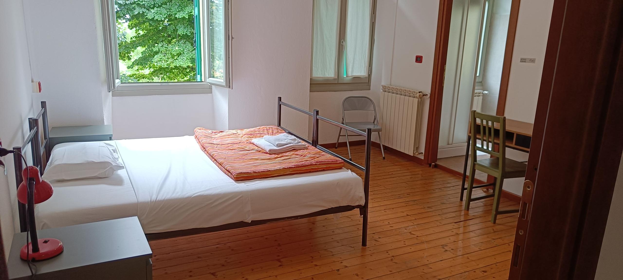 Bright private double room with wooden floors, a double bed with orange bedspread, desk, chair, and windows overlooking green trees