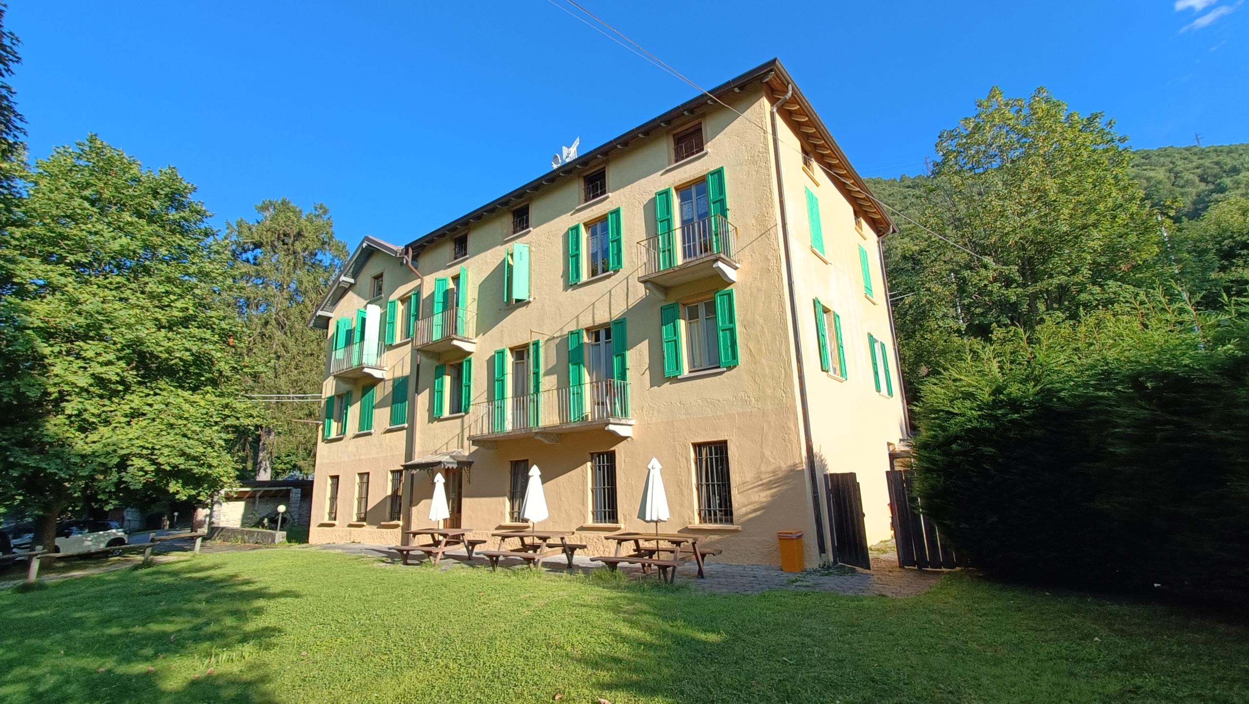 Exterior view of the Montino building with green shutters, surrounded by trees, with picnic tables and umbrellas in the garden