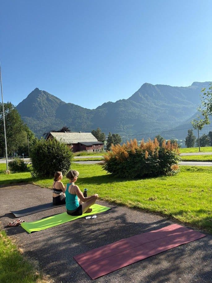 Two people doing yoga outdoors on mats with mountains and the Bjørkedal landscape in the background