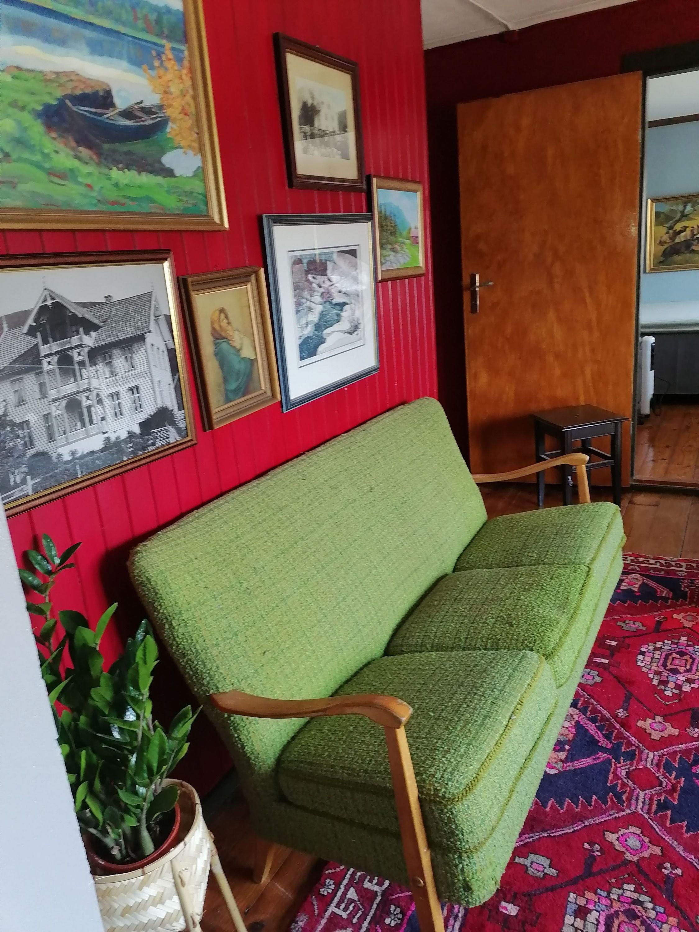 Lounge area with a green sofa, framed paintings on red walls, and a patterned rug