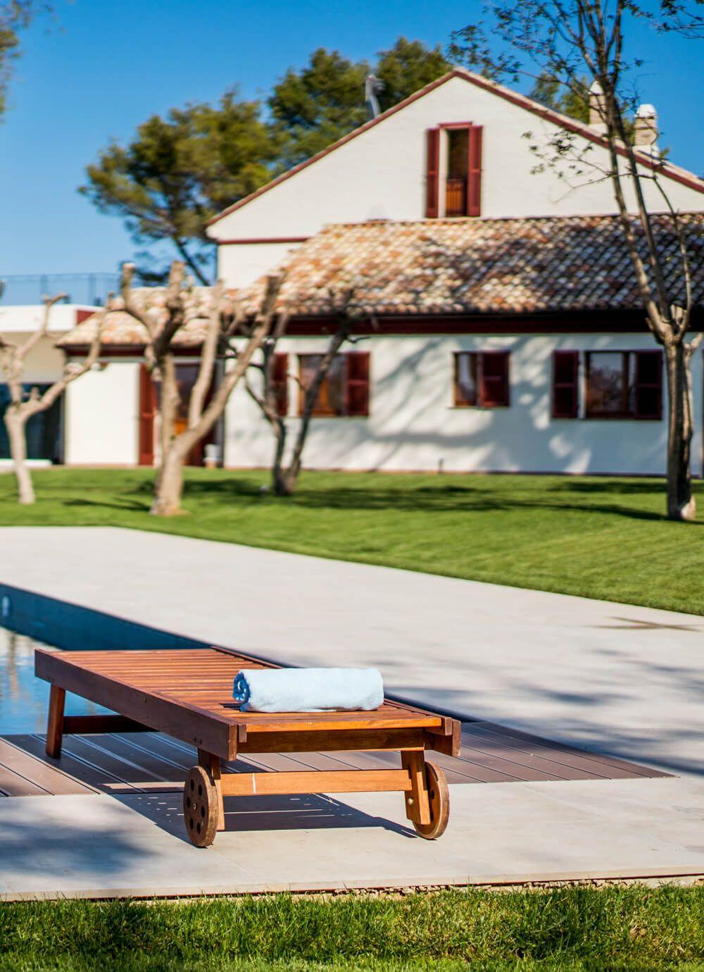 Wooden sun lounger with a rolled towel beside the pool, with the coliving country house building visible in the background