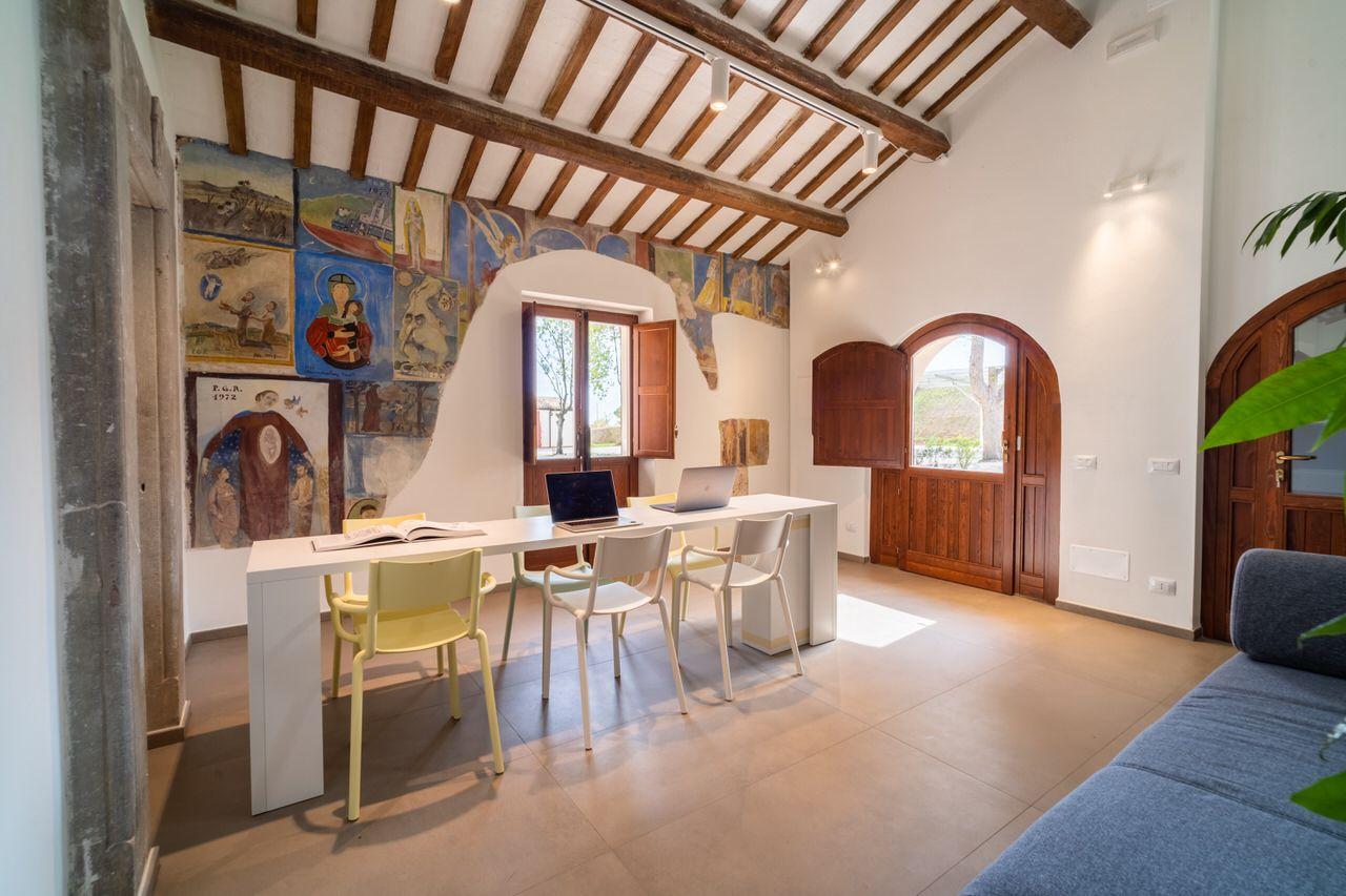 Room with frescoed walls, a long work/meeting table with laptops, chairs, and a private entrance with arched wooden doors