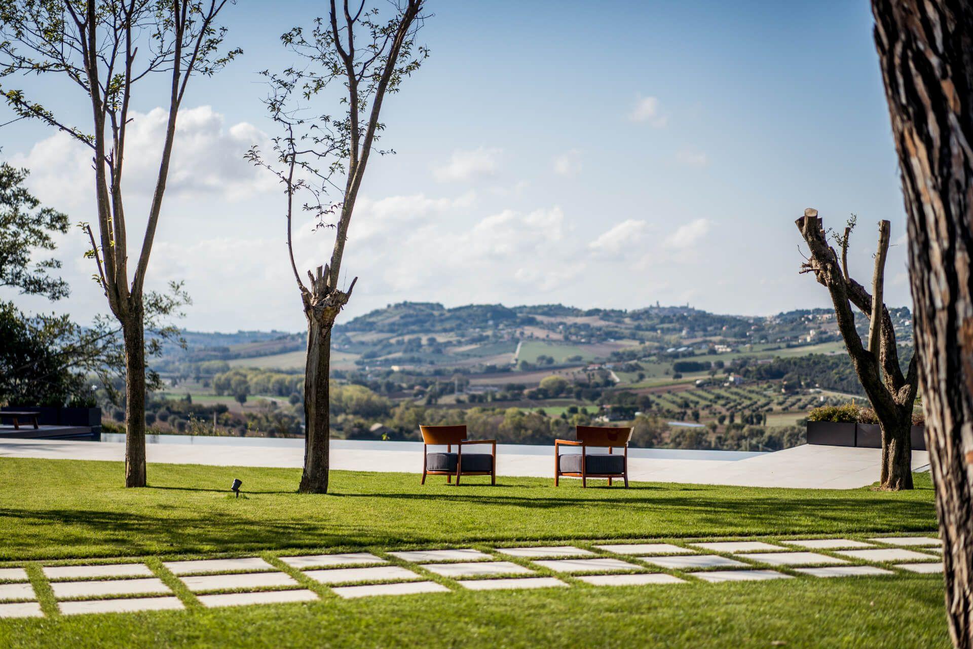 Outdoor garden with stepping stone path, trees, and two chairs beside the infinity pool overlooking the Marche hills