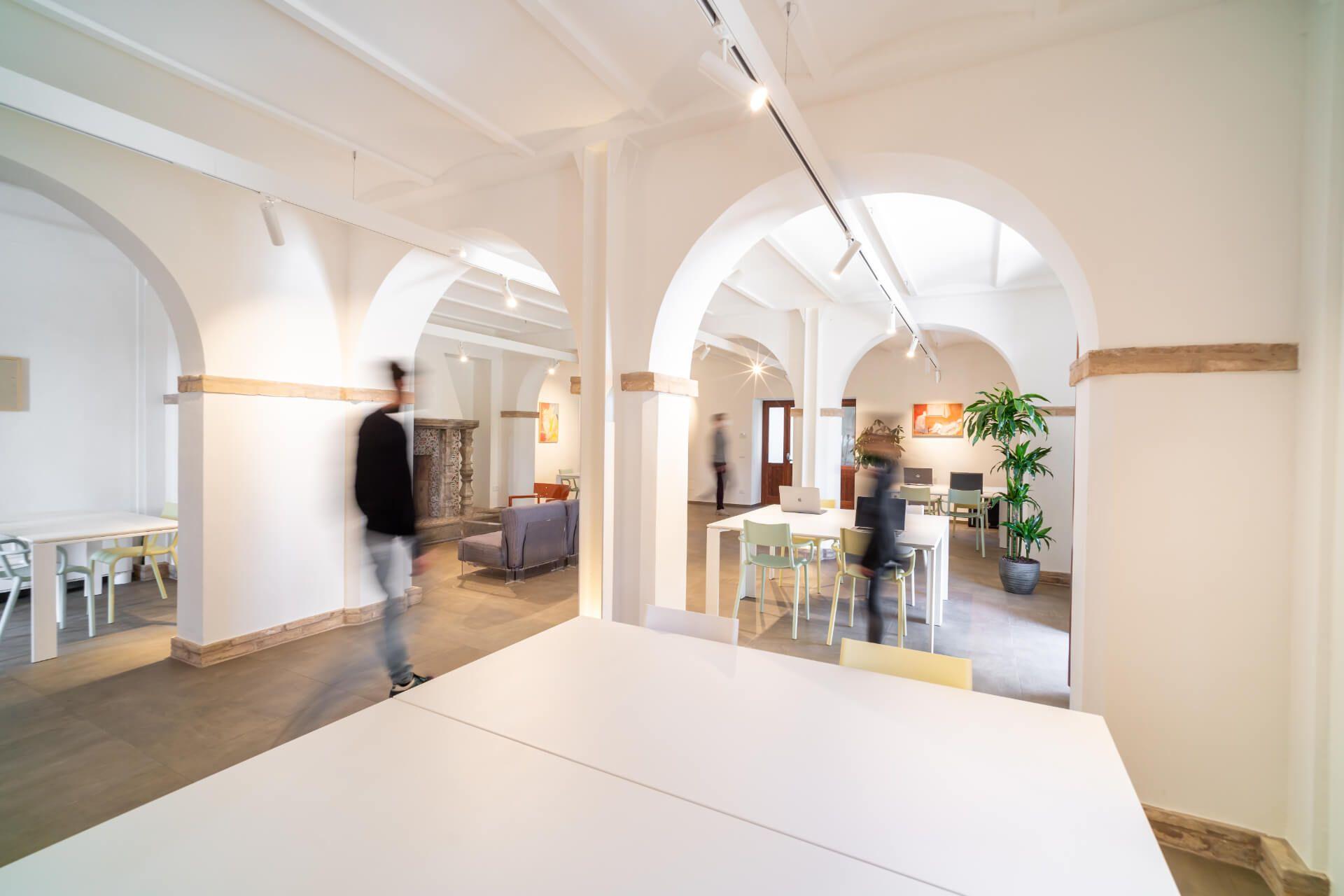 Open coworking space with white arched ceilings, wooden beams, work desks and a lounge area visible through the arches