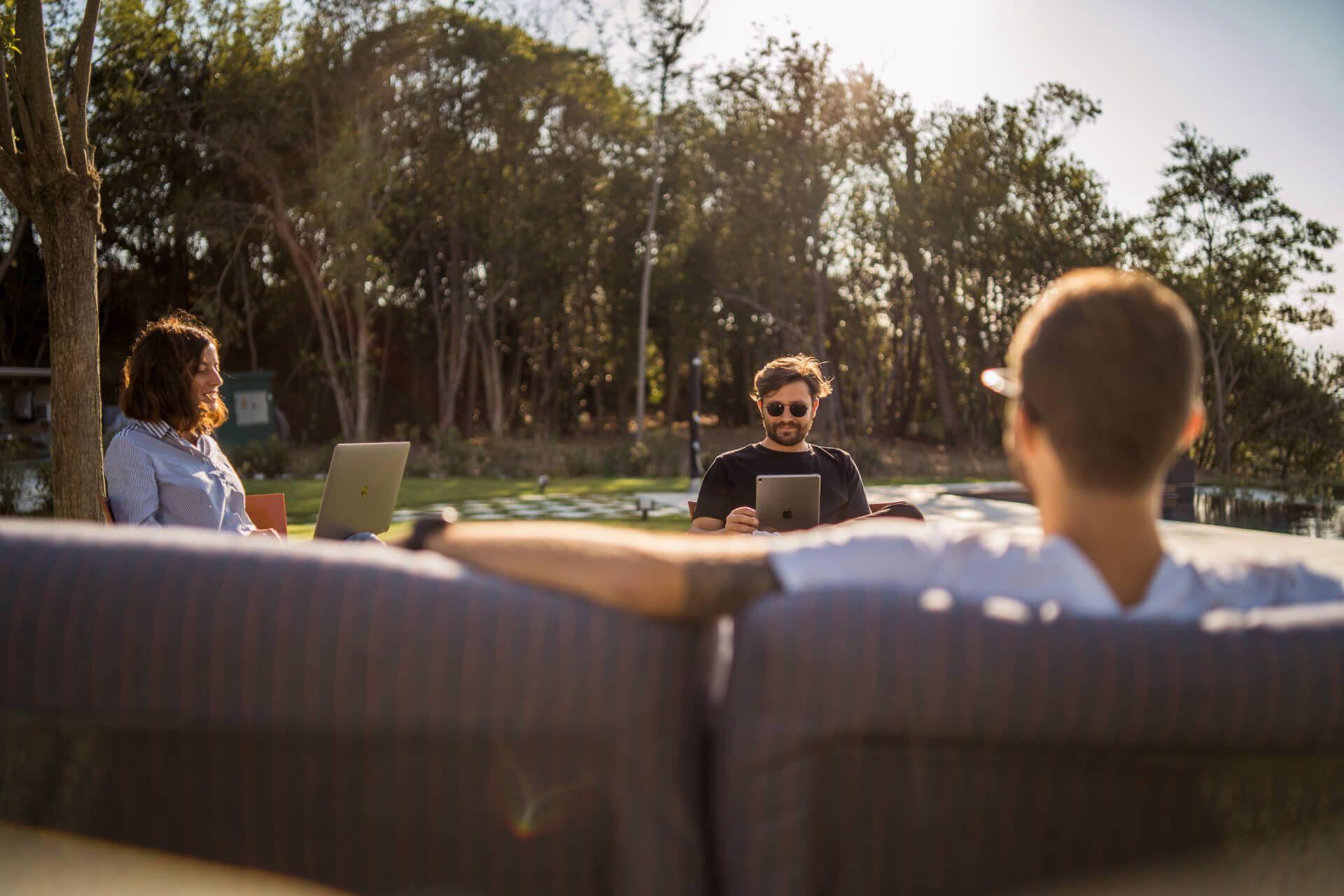 Three people relaxing and working outdoors on a sofa in the garden, with laptops and tablets, surrounded by trees
