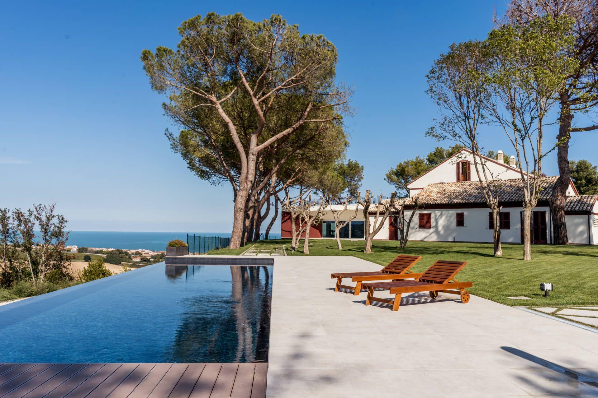 Outdoor infinity pool with sun loungers in the foreground and the coliving building visible behind trees, with a view of the Adriatic Sea in the background