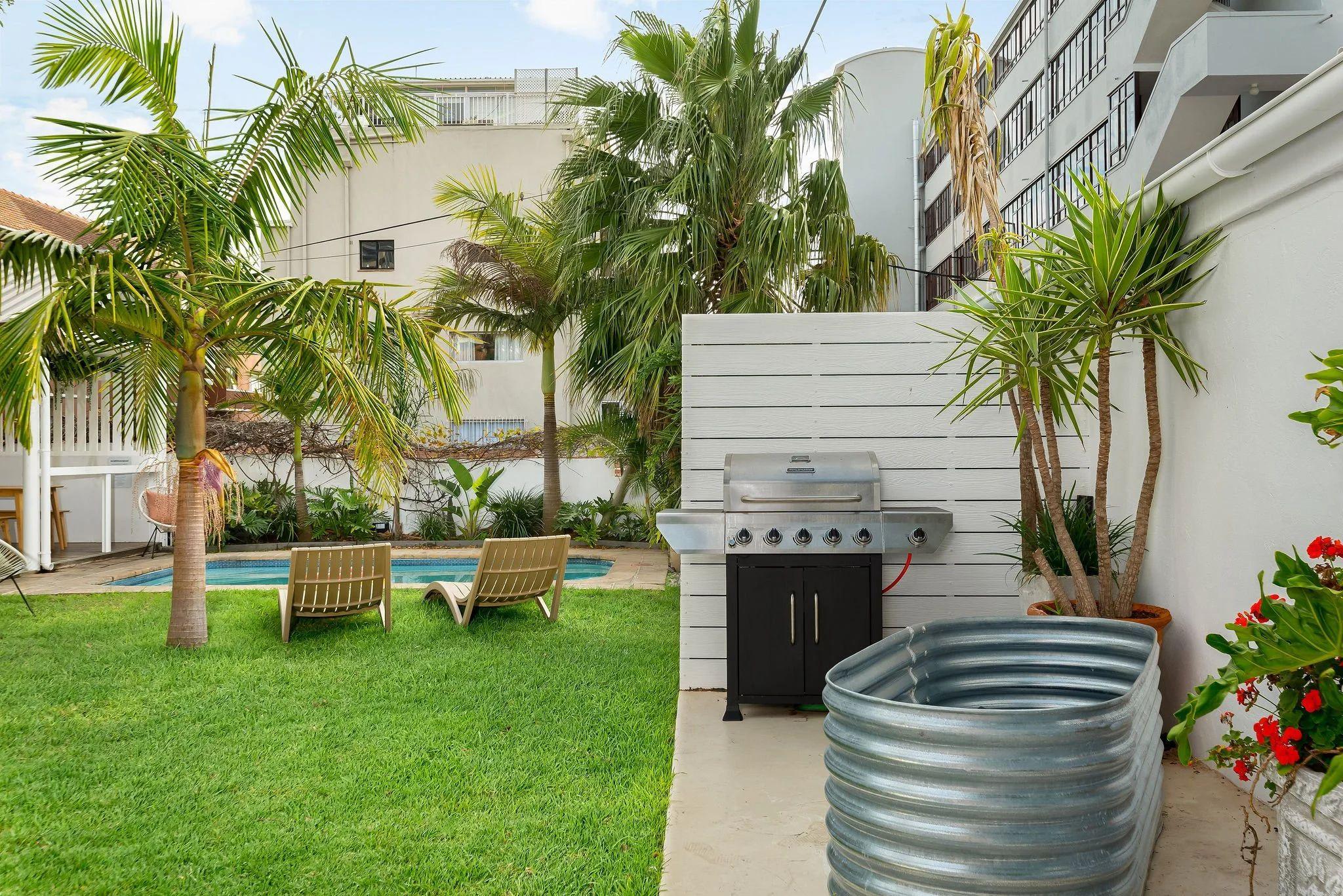 Lush garden at Neighbourgood Hill with outdoor pool visible in the background, gas BBQ grill, sun loungers, and palm trees