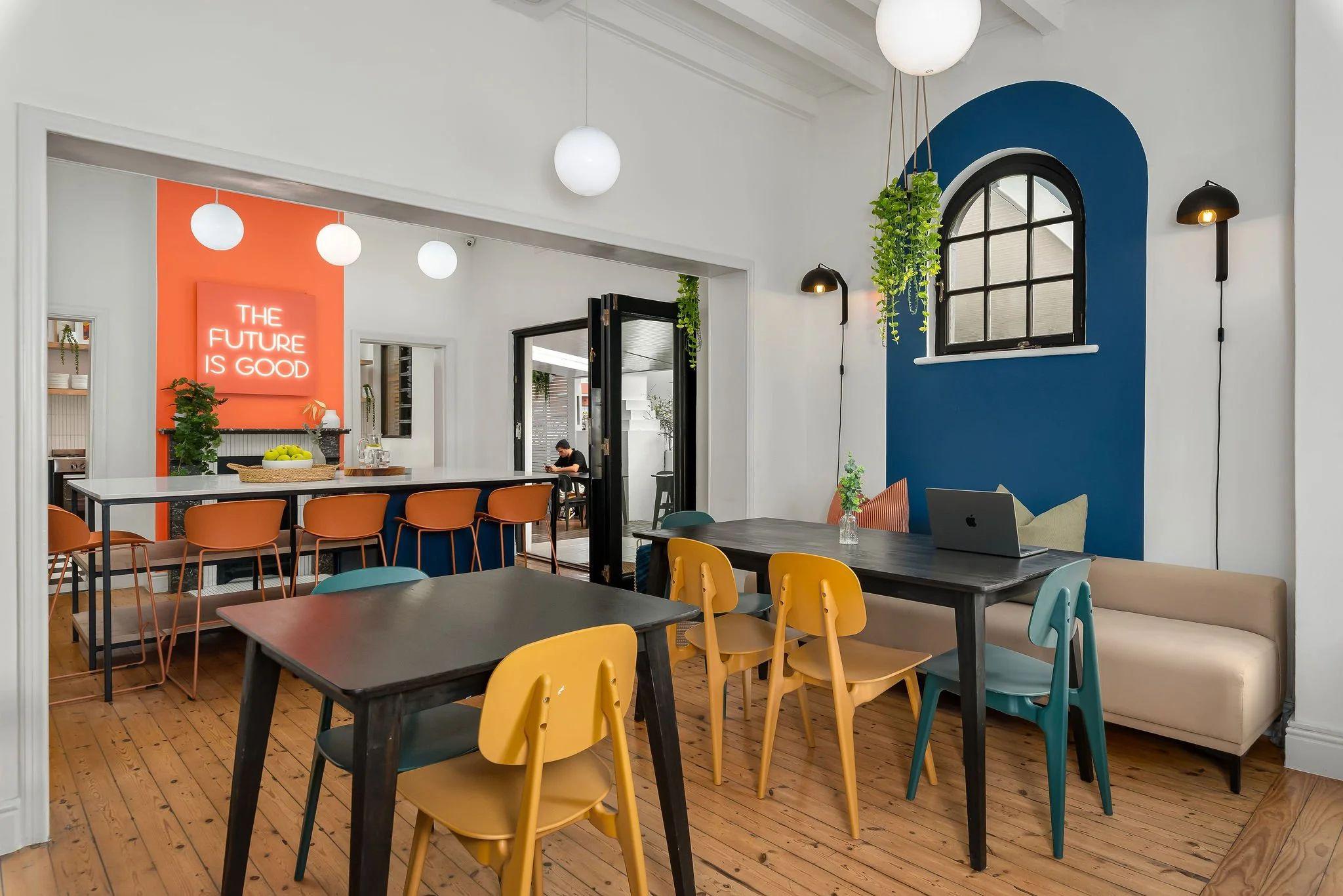 Open-plan workspace lounge at Neighbourgood Hill with colourful chairs, work tables, and neon 'The Future Is Good' sign visible in the background
