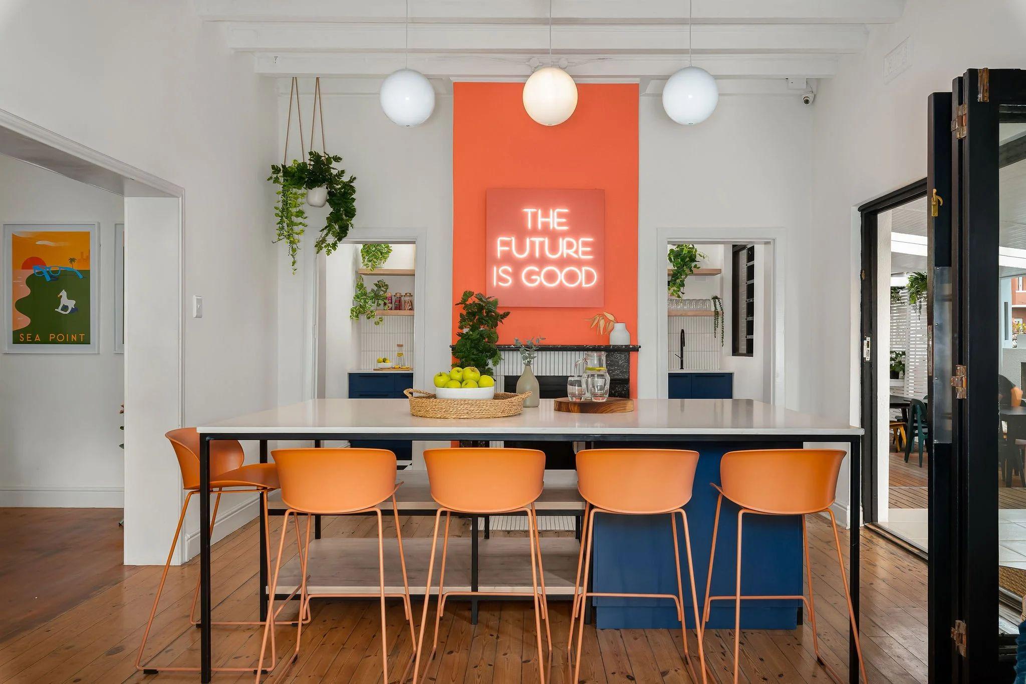 Shared kitchen area at Neighbourgood Hill featuring a neon 'The Future Is Good' sign, island counter with orange bar stools, and open plan layout