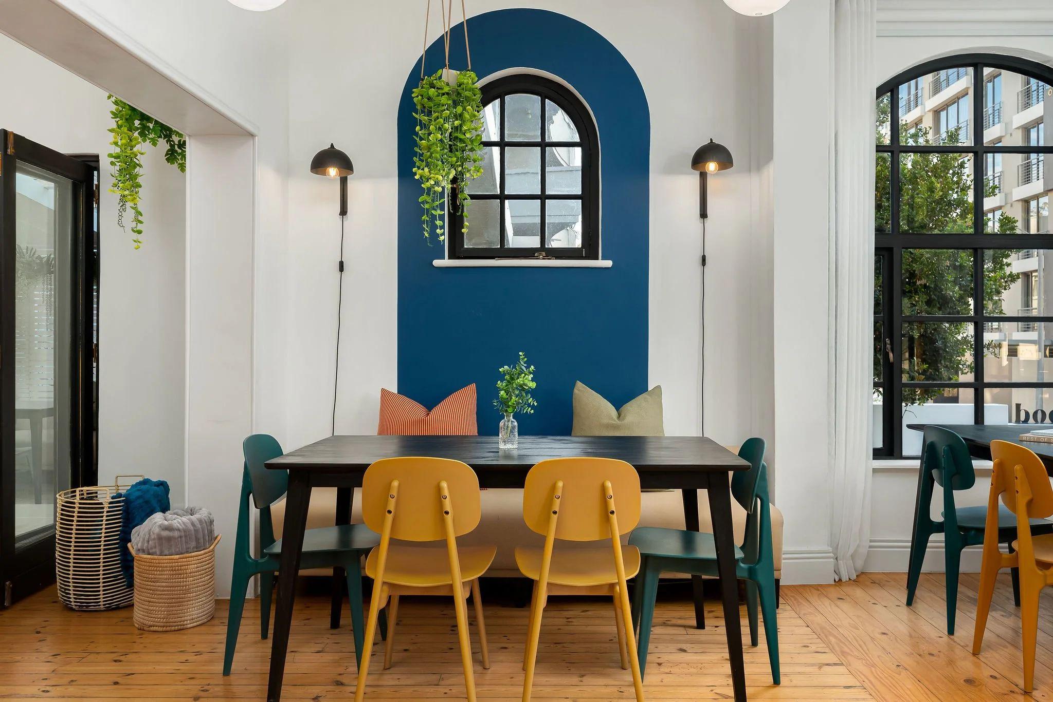 Dining and workspace area at Neighbourgood Hill with dark tables, yellow and teal chairs, and blue arched wall feature