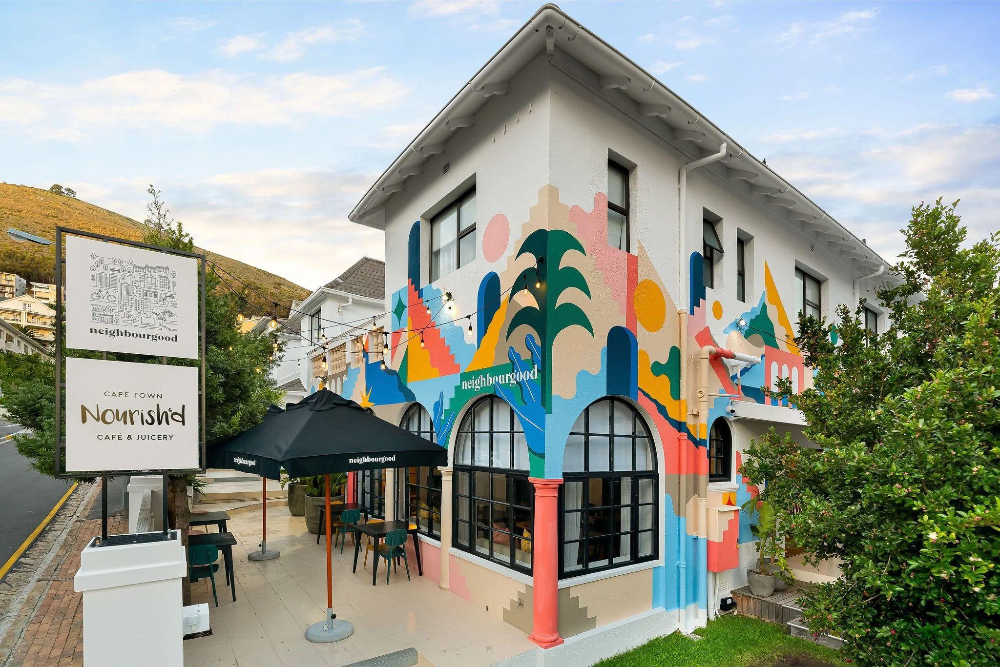 Exterior view of Neighbourgood Hill building in Green Point, featuring the colourful mural facade and Nourish'd Café & Juicery signage with outdoor seating