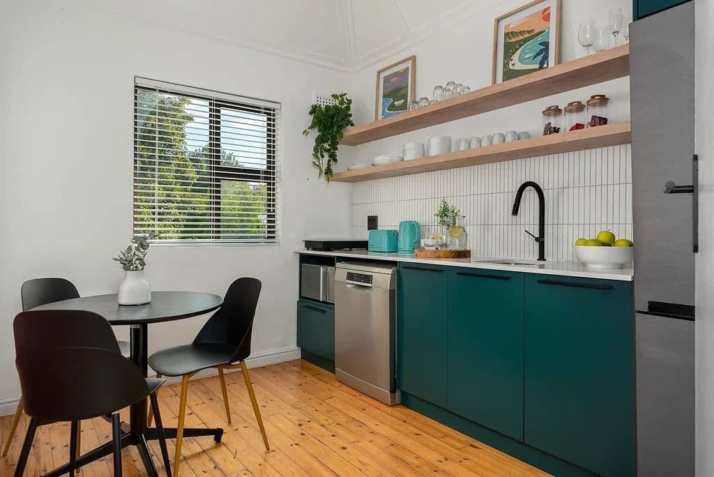 Kitchen in the two-bedroom apartment at Neighbourgood Hill with teal cabinetry, open shelving, dishwasher, and small dining table