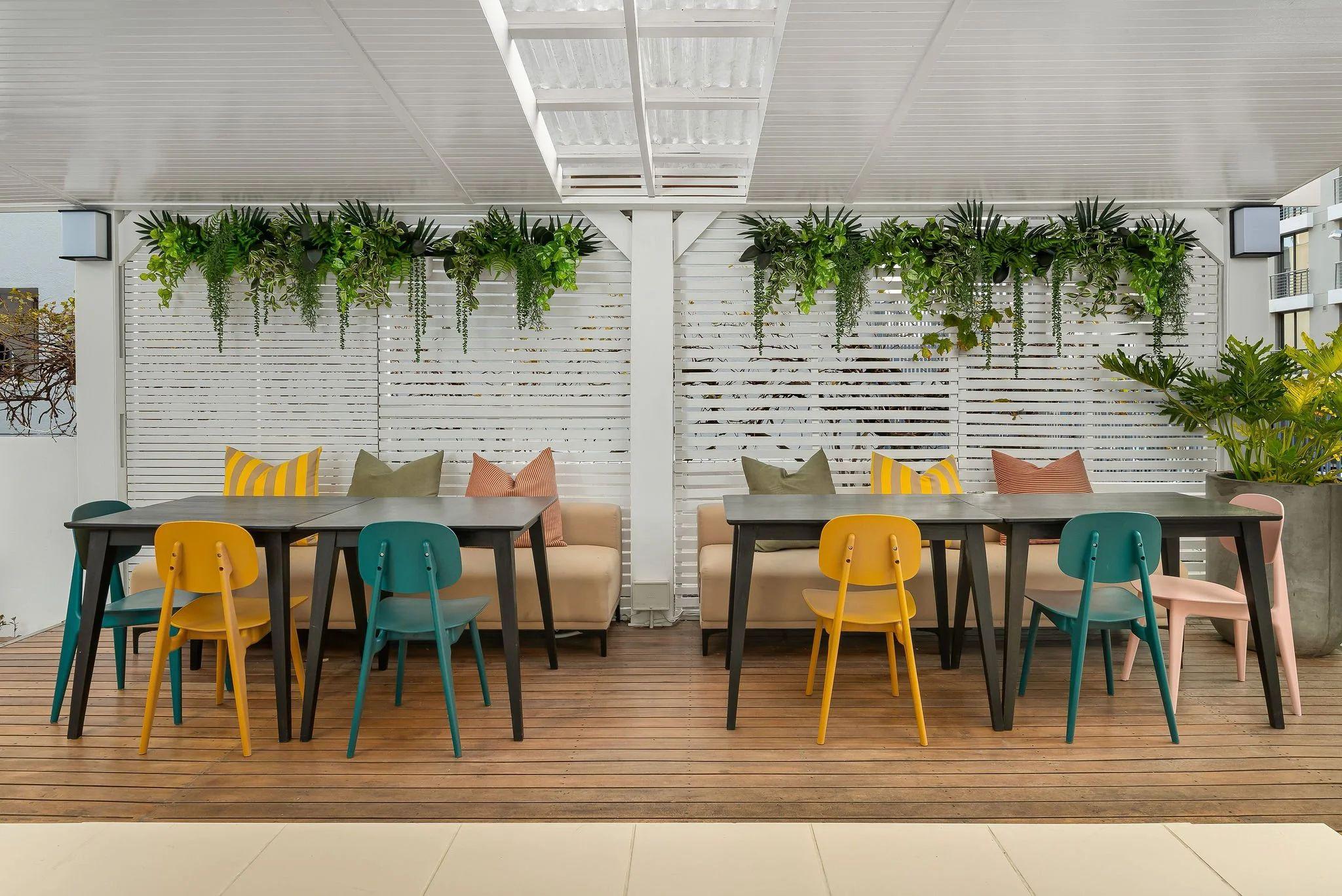 Covered terrace seating area of Nourish'd Café at Neighbourgood Hill with colourful chairs and hanging plants
