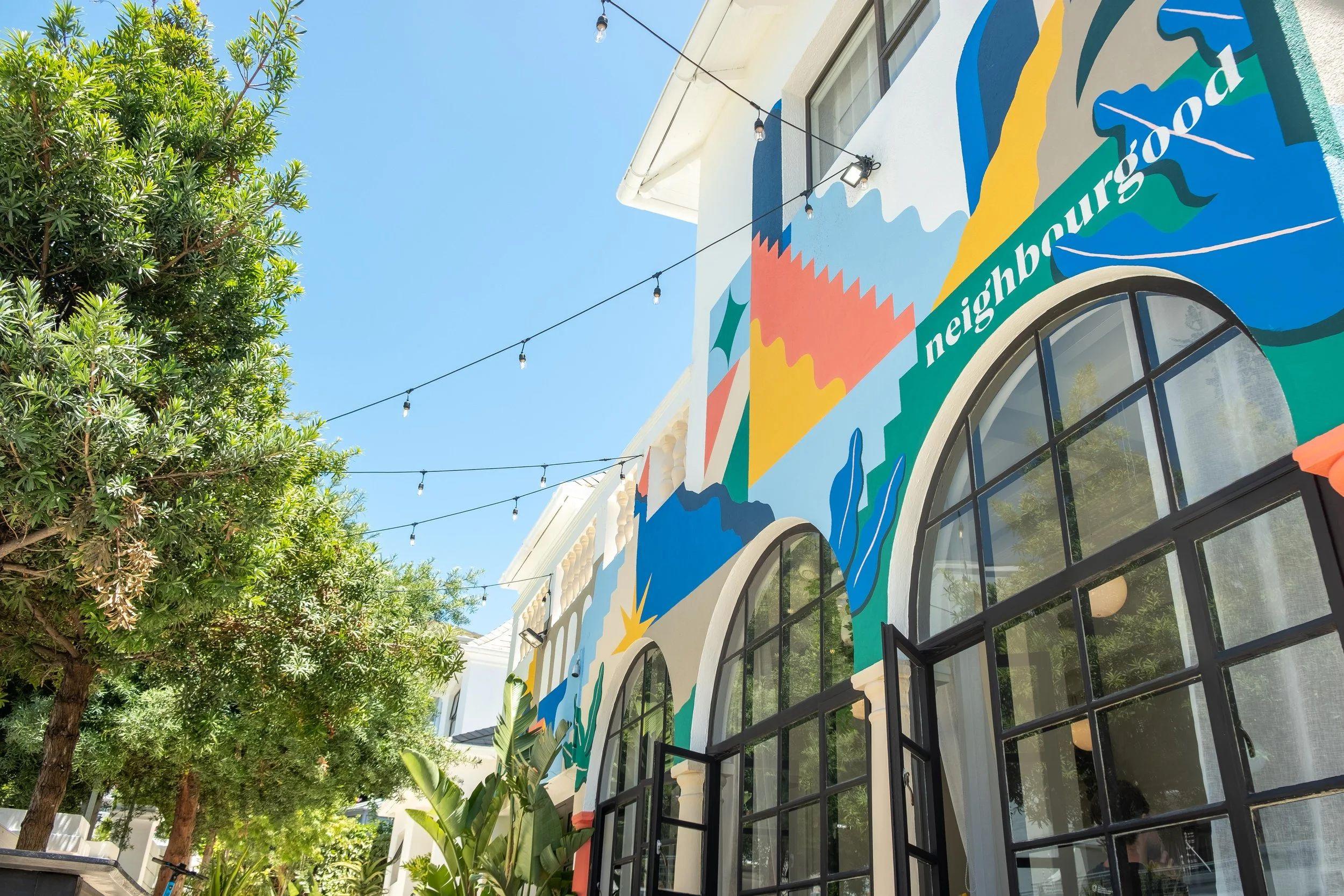 Close-up view of the colourful Neighbourgood mural on the exterior wall of Neighbourgood Hill, with string lights and arched windows