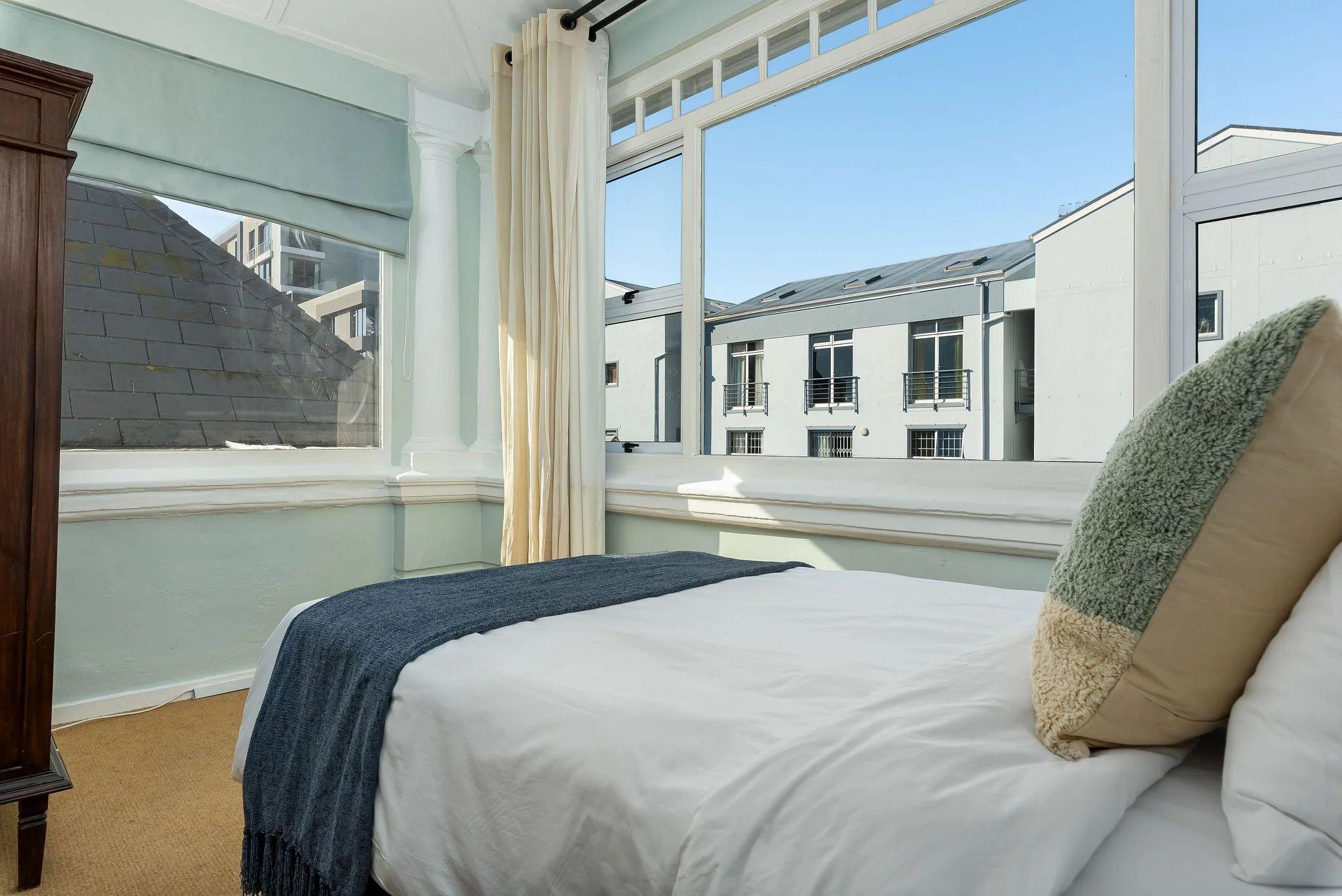 Bright Hill Suites room with large windows overlooking the neighbourhood, white bedding with blue throw, and green headboard