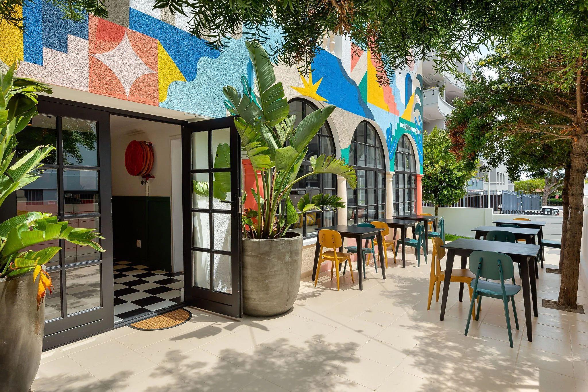 Outdoor terrace seating area of Nourish'd Café at Neighbourgood Hill, with colourful mural on the building wall and tables with chairs
