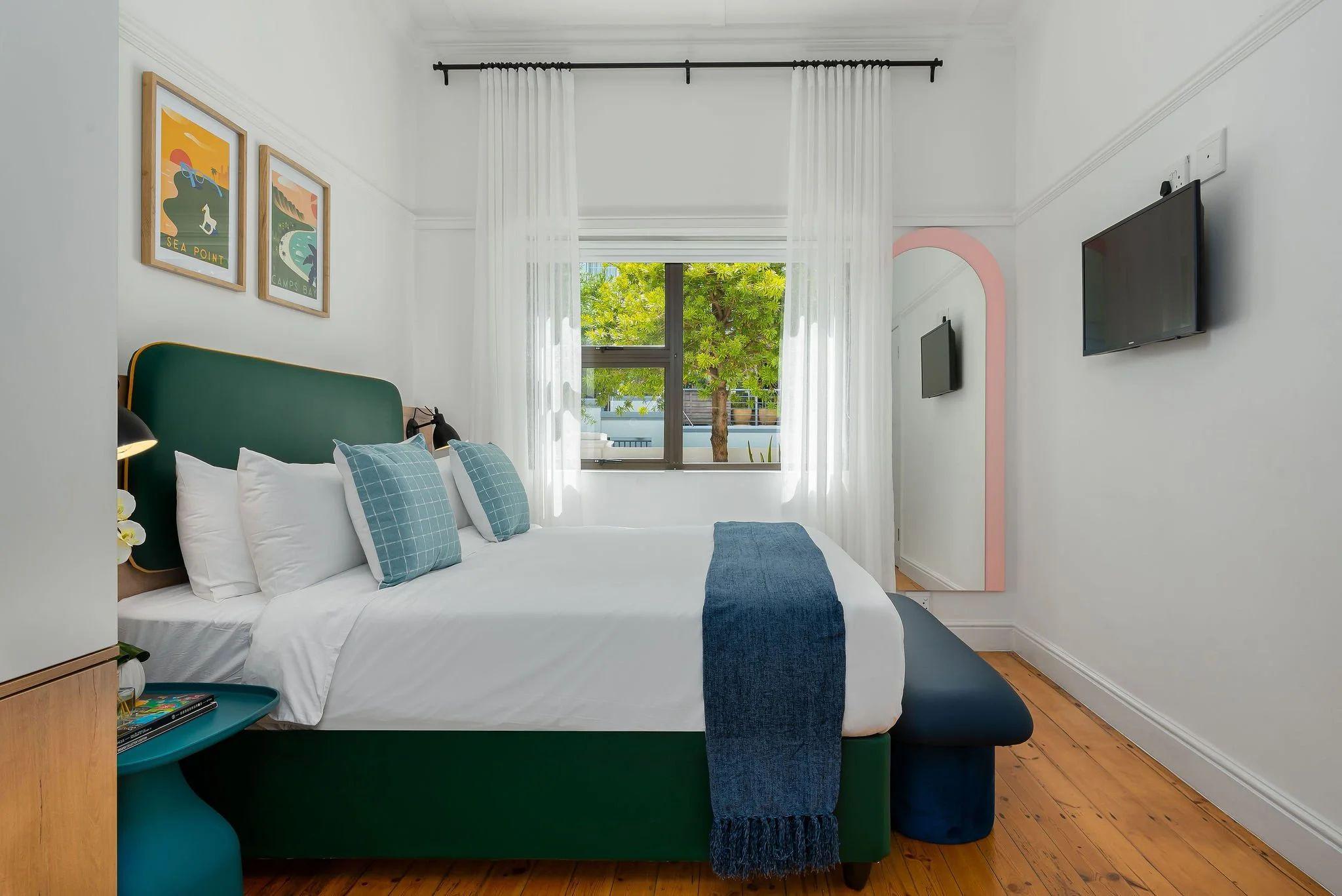 Standard room at Neighbourgood Hill with green upholstered headboard, blue bench, travel posters, and flat-screen TV