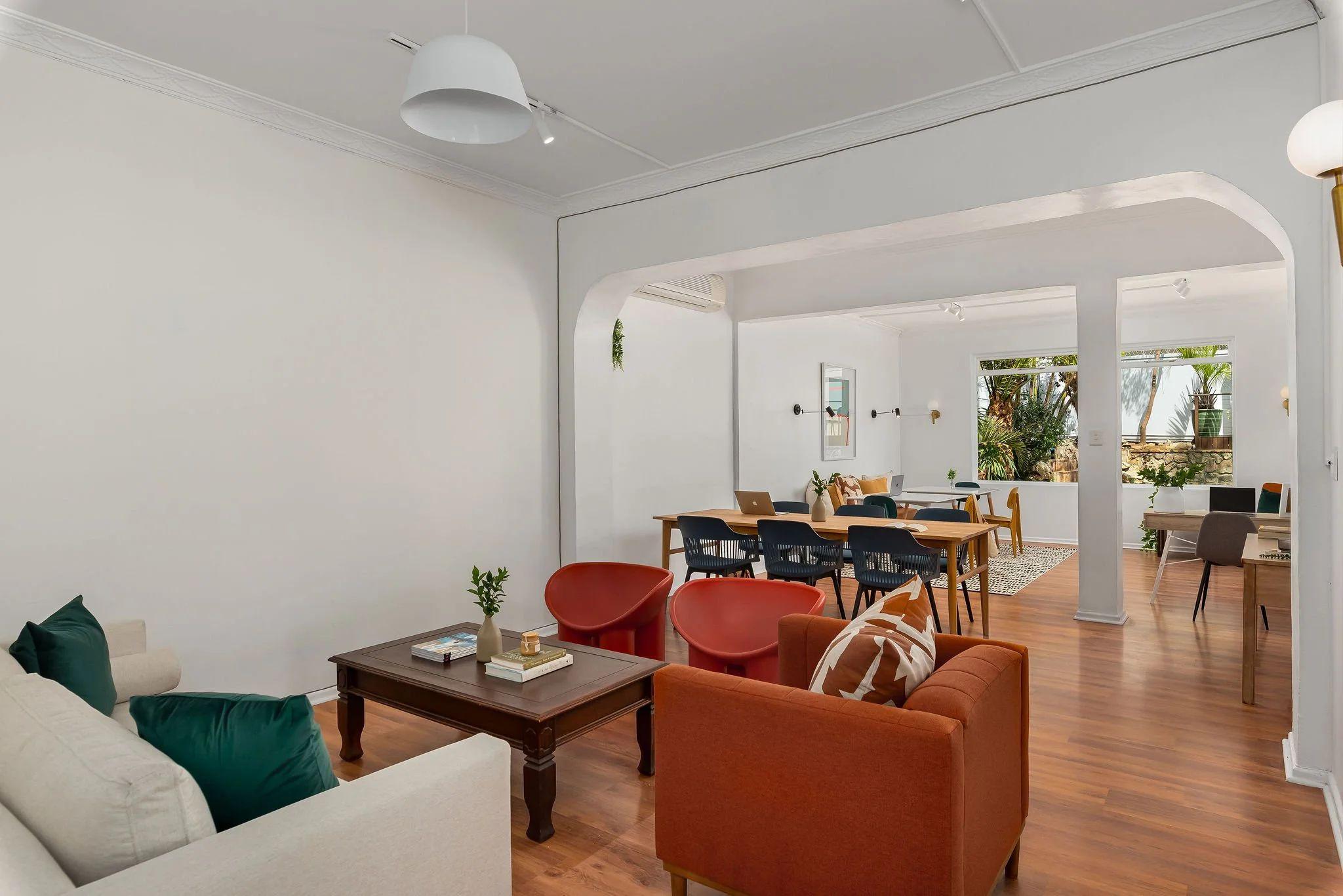 Open-plan communal lounge with sofas, armchairs, and a dining/workspace area visible through an archway, with wooden floors
