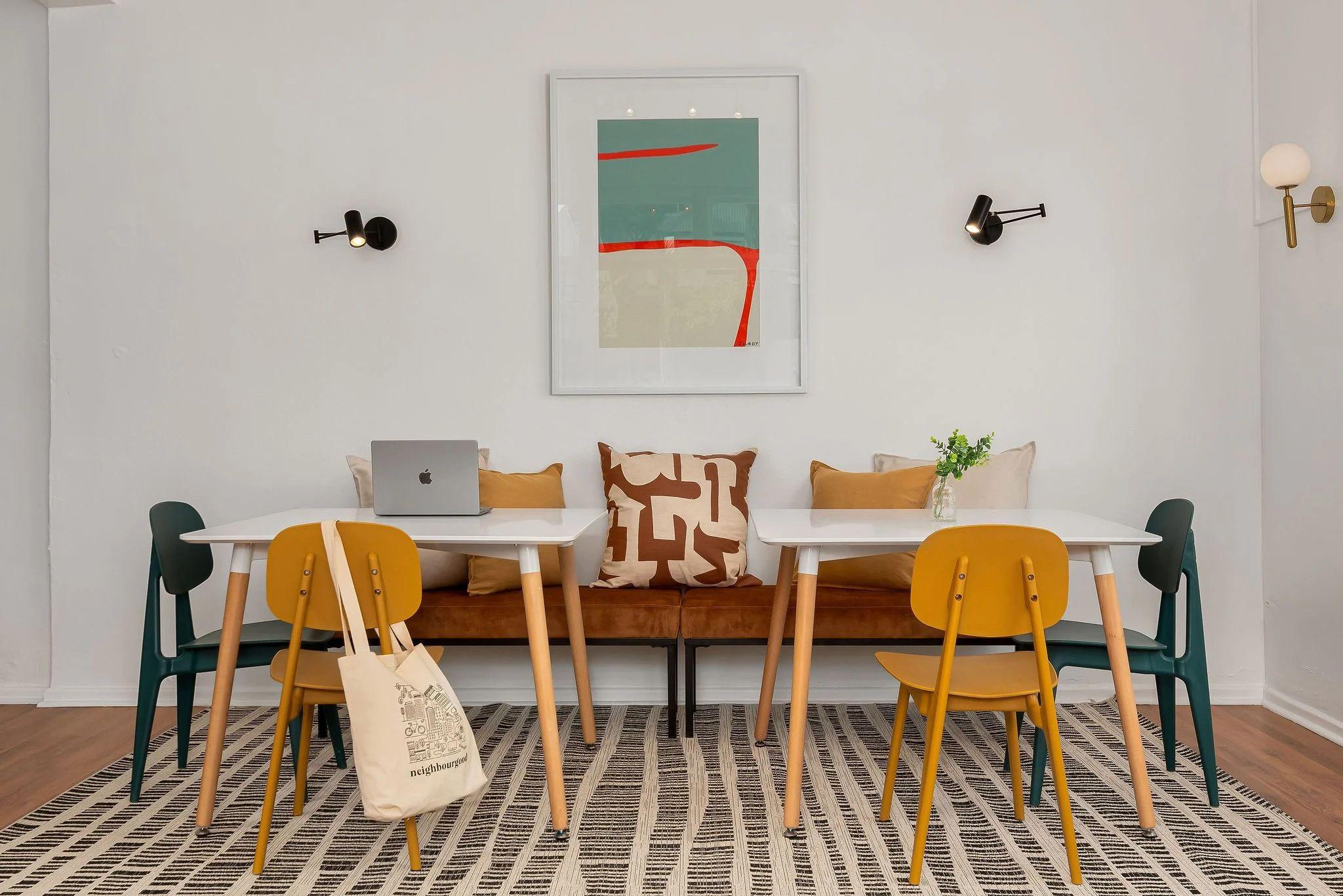 Communal workspace with a white dining table, yellow and teal chairs, bench seating, and a laptop on the table