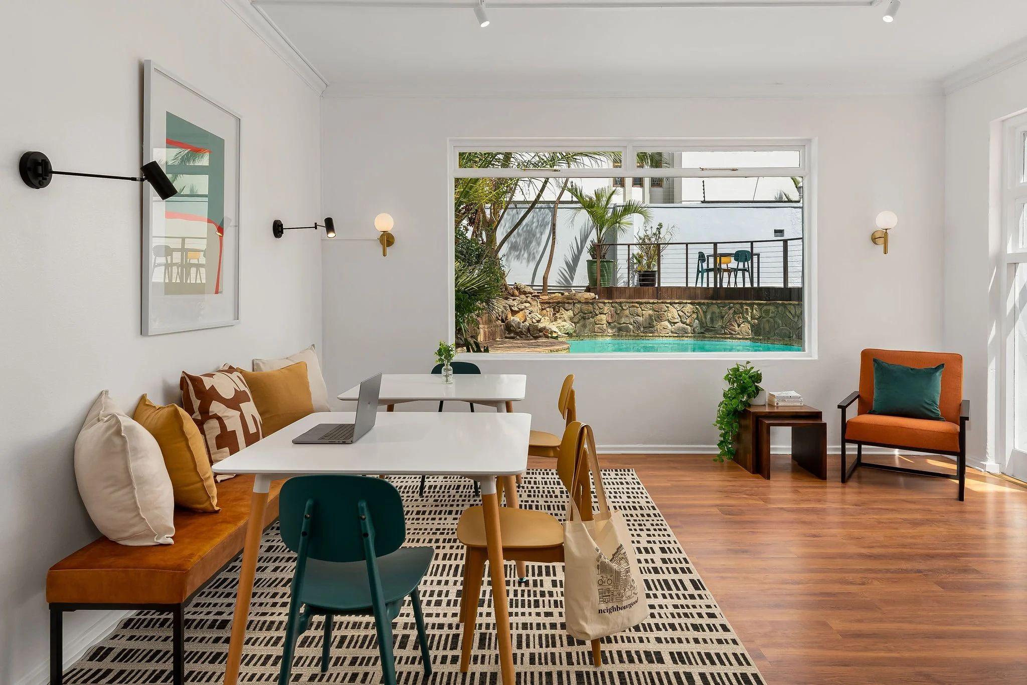 Communal workspace and dining area with a large window overlooking the outdoor pool, featuring a white table, colourful chairs, bench seating with cushions, and an armchair