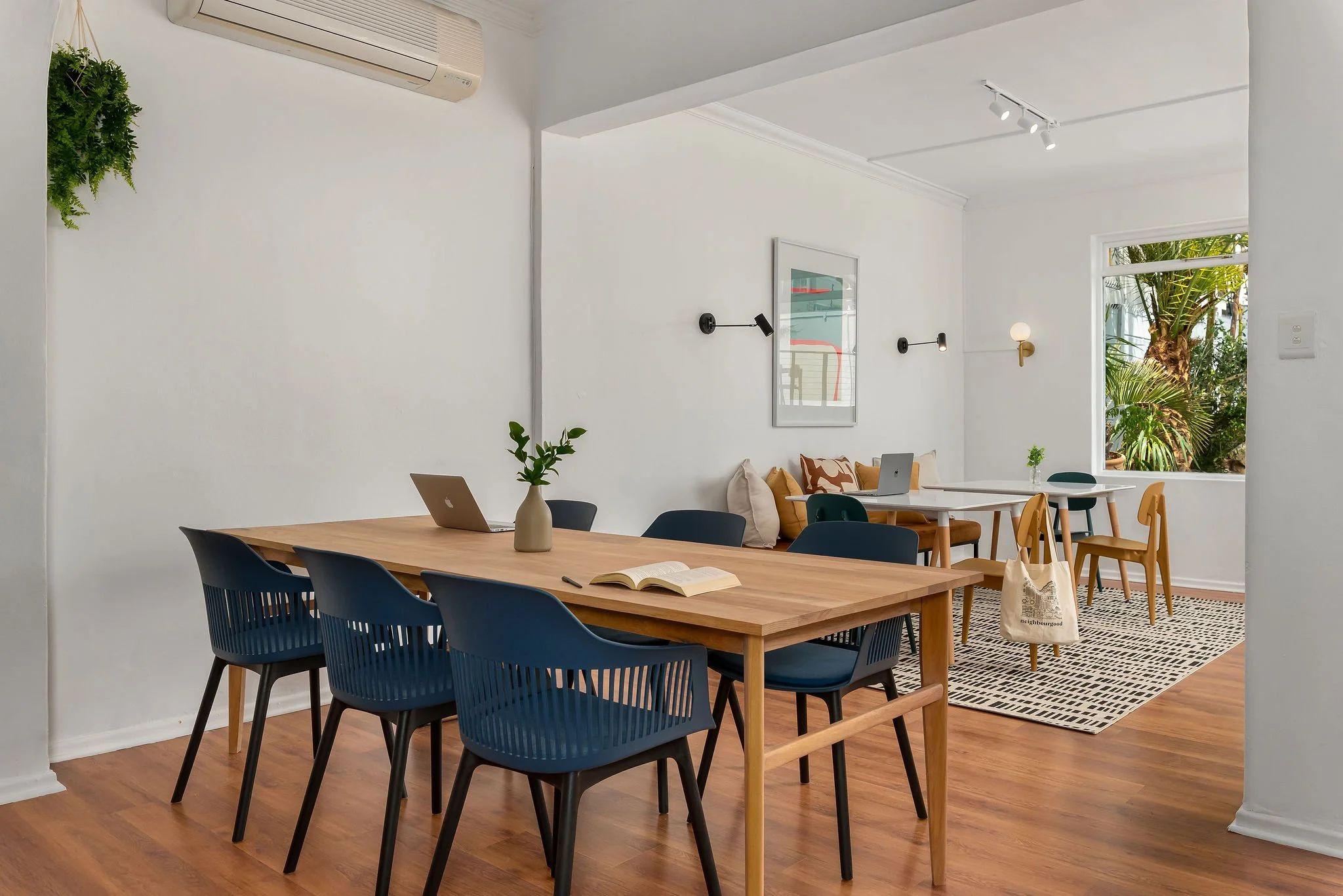 Communal workspace with a large wooden table, navy blue chairs, laptop, and open book, with the lounge area visible in the background