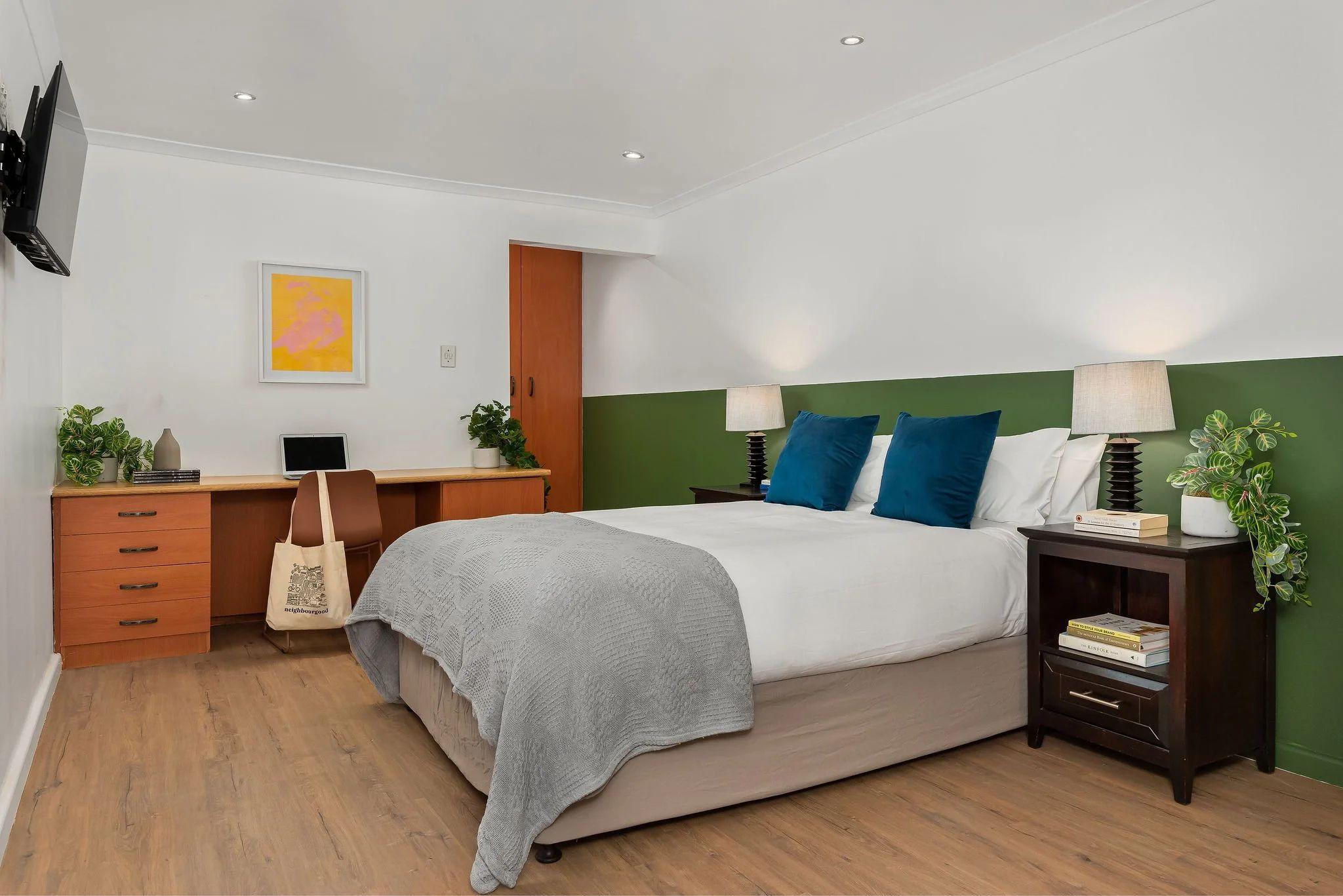 Deluxe room with a queen-size bed, blue cushions, green accent wall, wooden desk with laptop, and nightstands with lamps