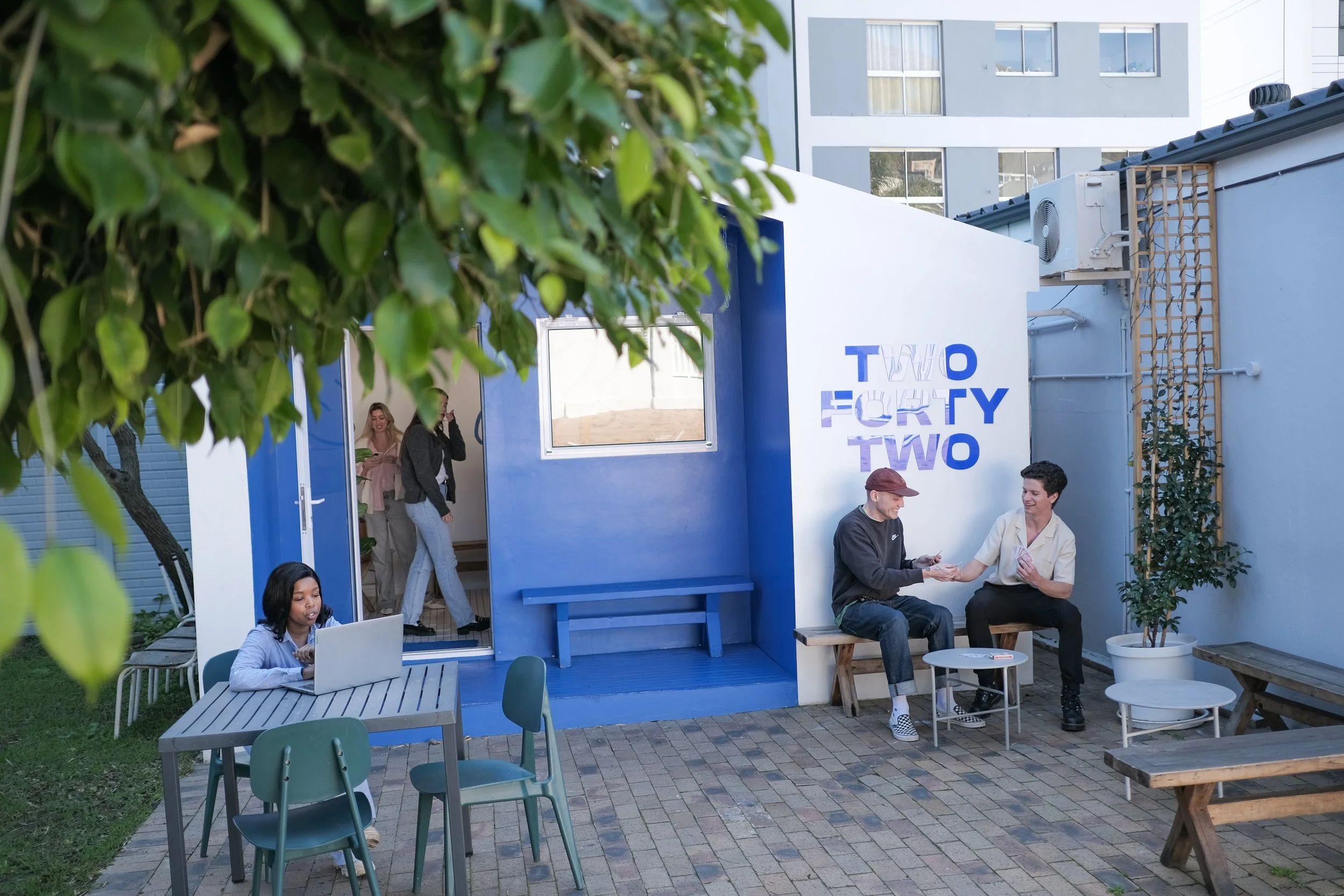 Outdoor courtyard area with a person working on a laptop at a table, and two others chatting near a blue-painted wall with 'Two Forty Two' signage