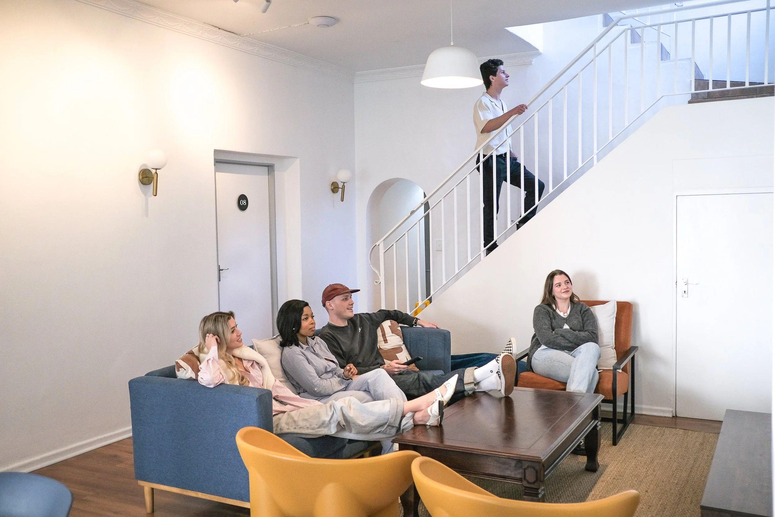 Group of guests socialising in the communal lounge area with sofas, armchairs, and a staircase in the background