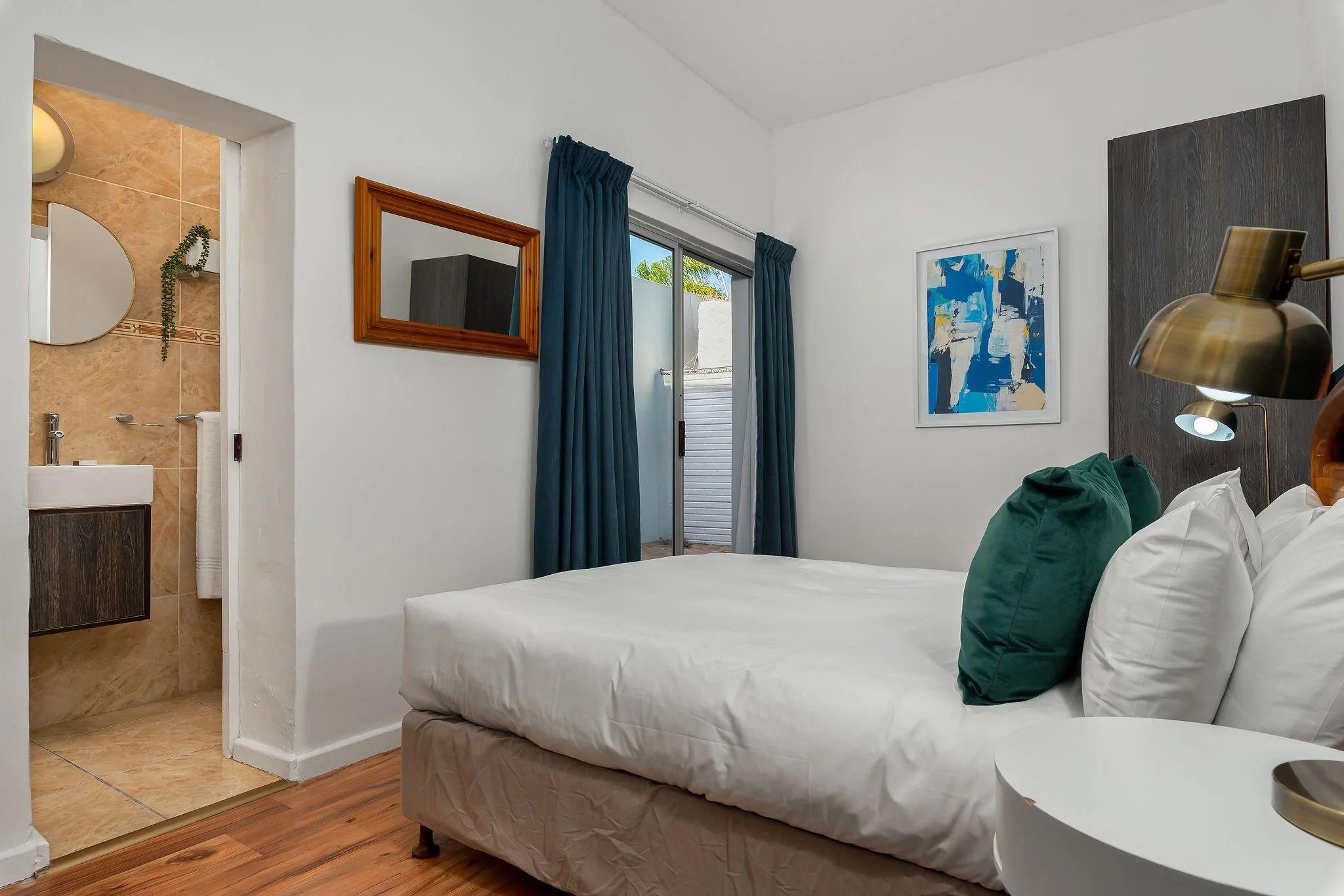 Standard room with a double bed, green cushions, en-suite bathroom visible, sliding door to balcony, and wall-mounted lamp