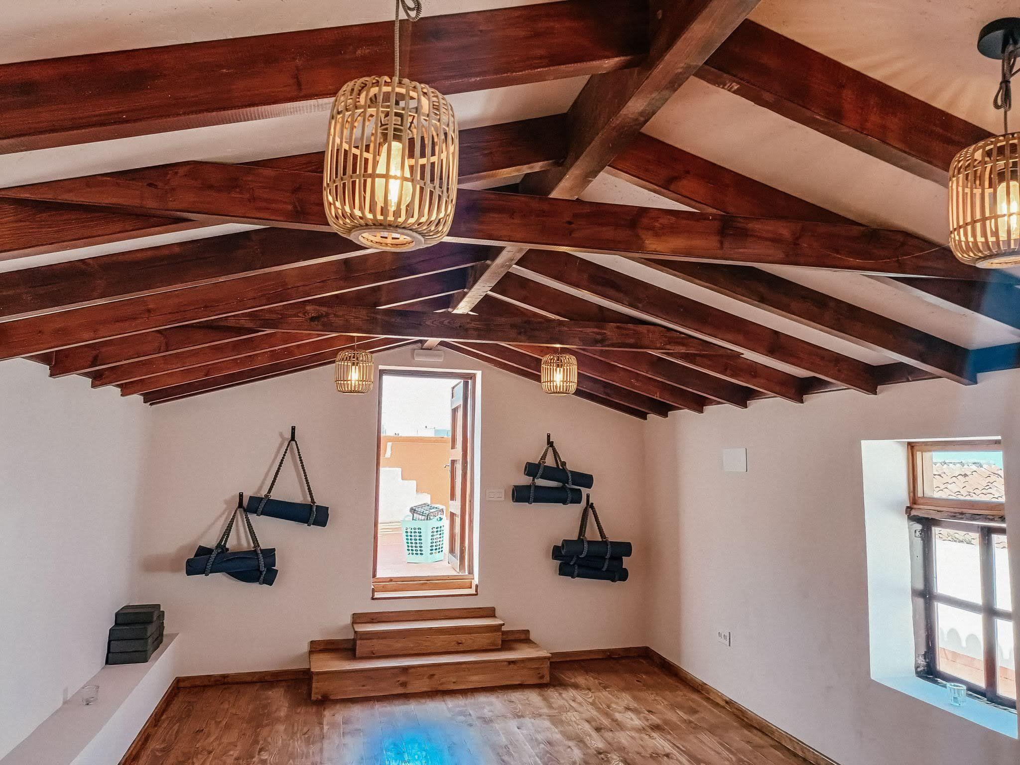 The yoga shala at Nine Coliving with exposed wooden beam ceiling, rattan pendant lights, yoga mats hanging on the walls, and a raised wooden platform.