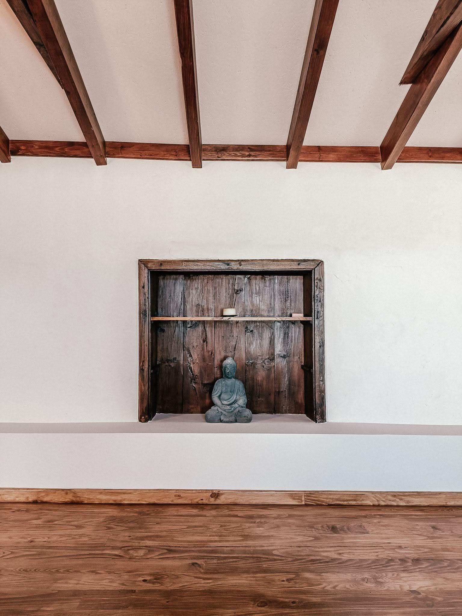 Detail of the yoga shala interior showing a Buddha statue in a wooden niche, exposed beam ceiling, and wooden floor.