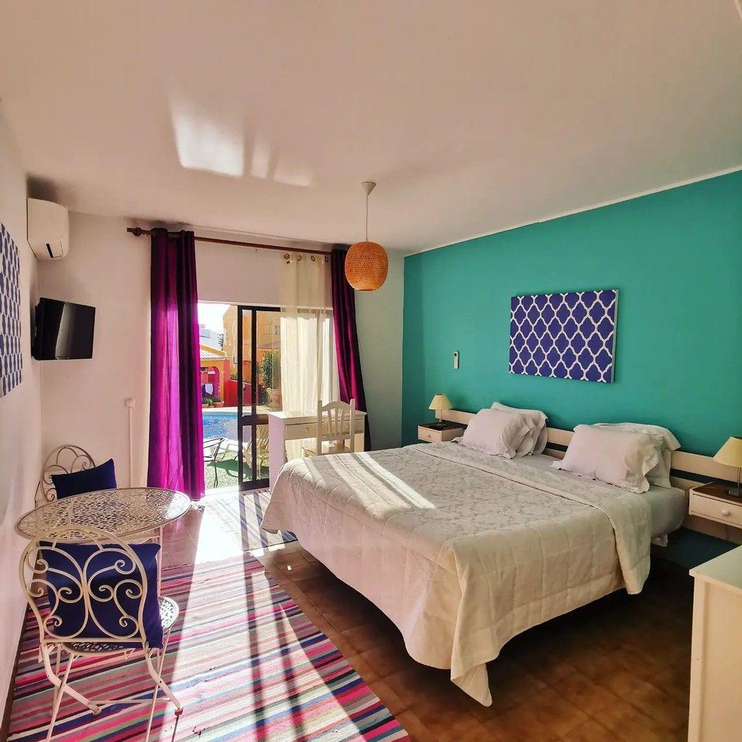 Bright private room with teal accent wall, queen bed, work desk, TV and balcony access to pool