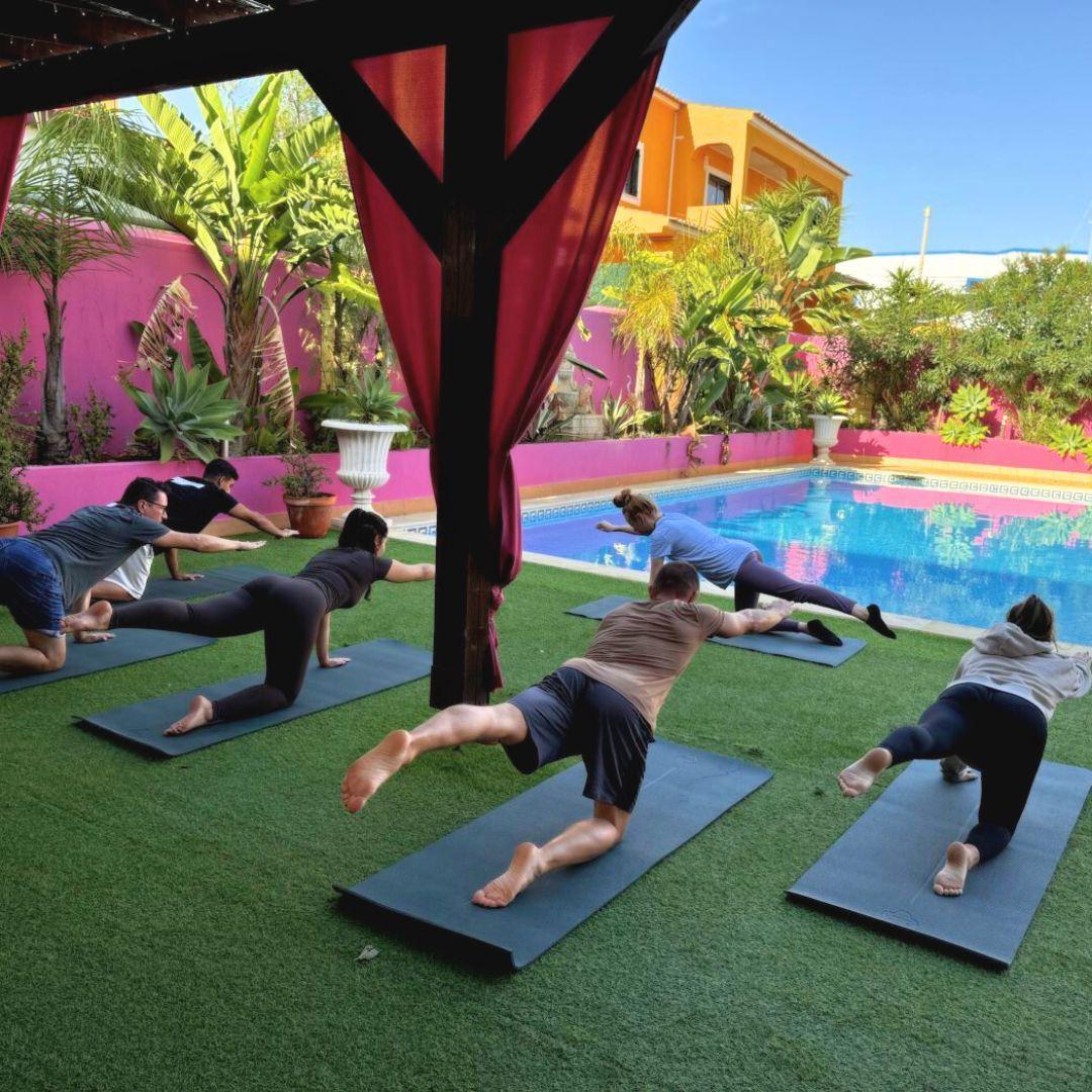 Community members doing yoga on mats by the pool under a pergola with pink curtains