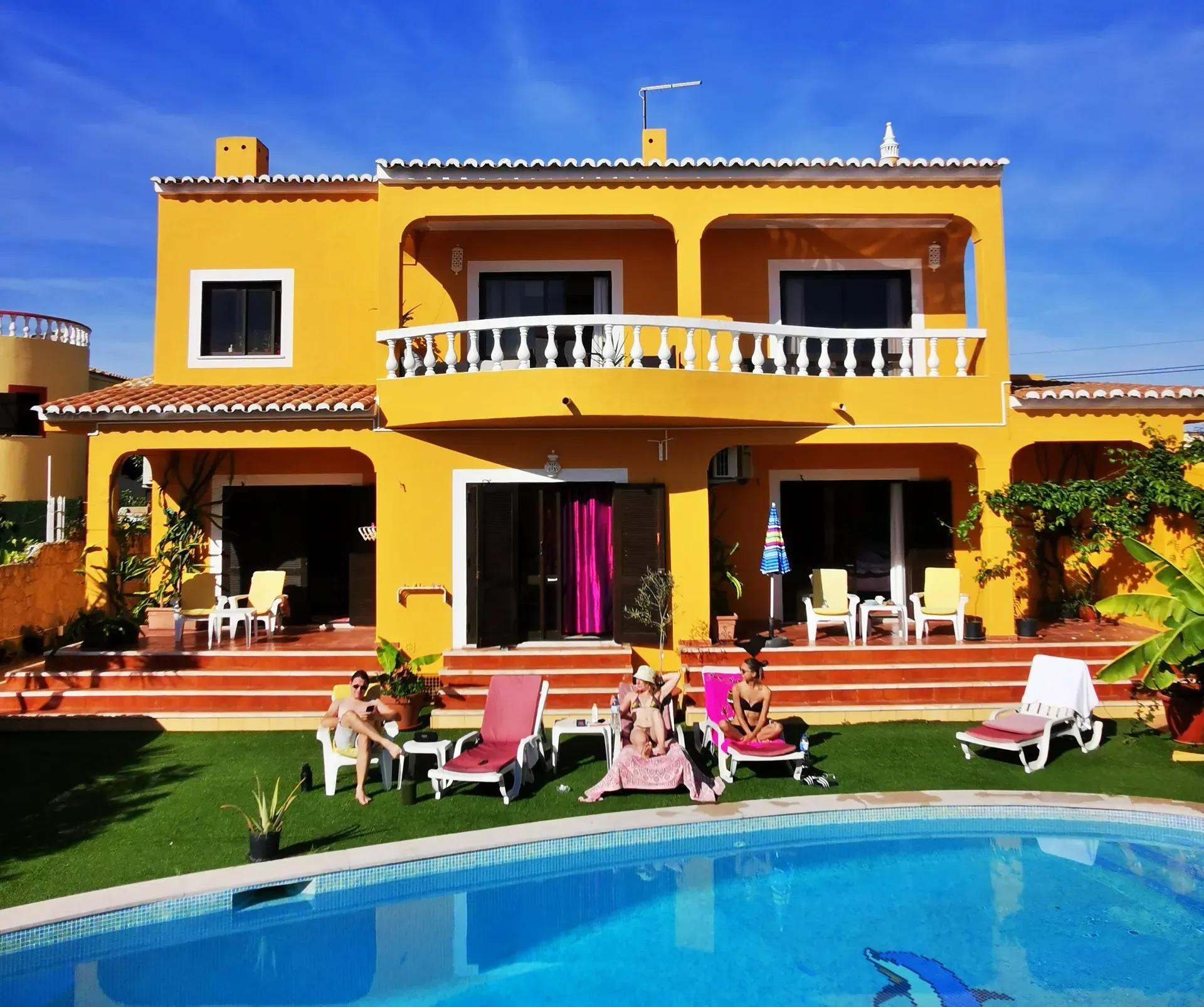 Yellow villa exterior with swimming pool and guests relaxing on sun loungers