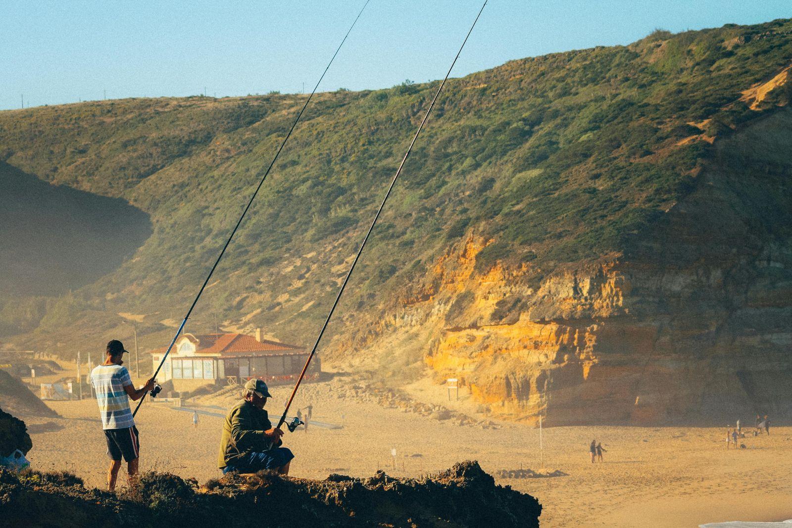 Two fishermen on rocks at a sandy beach near Ericeira with cliffs in the background