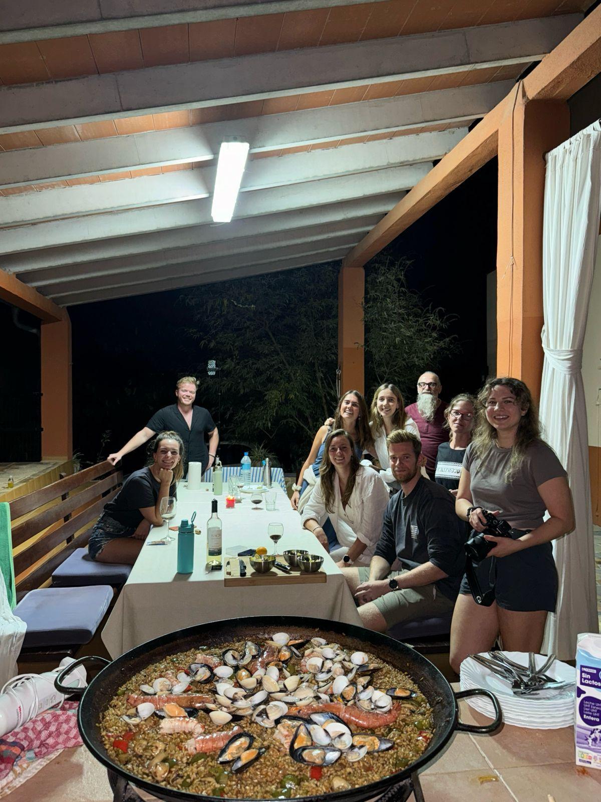 Group of guests gathered around a large paella pan for a community dinner on the covered patio
